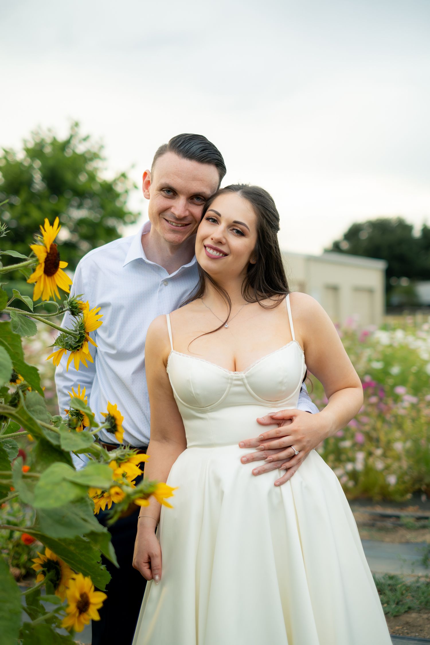Smiling couple posing by sunflowers, bride in white bustier dress with groom in white shirt in a flower garden.