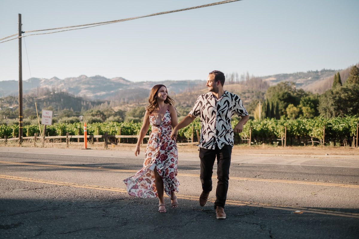 Couple walking hand in hand on a sunny road with vineyard and mountains in background.