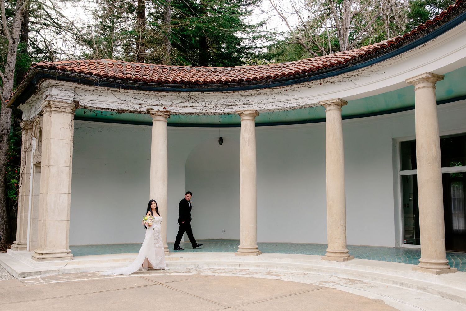 Bride and groom walking under elegant curved colonnade with white columns at an outdoor wedding venue.
