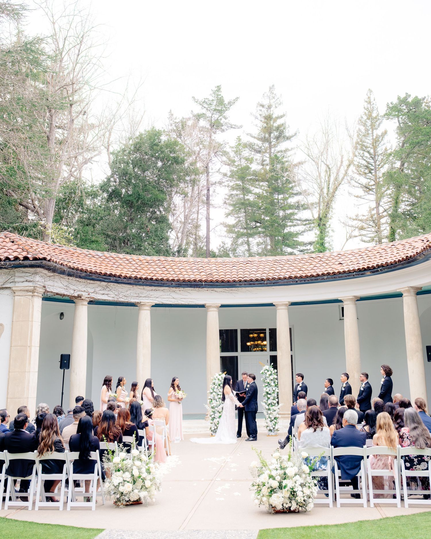 Elegant outdoor wedding ceremony at columned venue with white floral arrangements and seated guests.