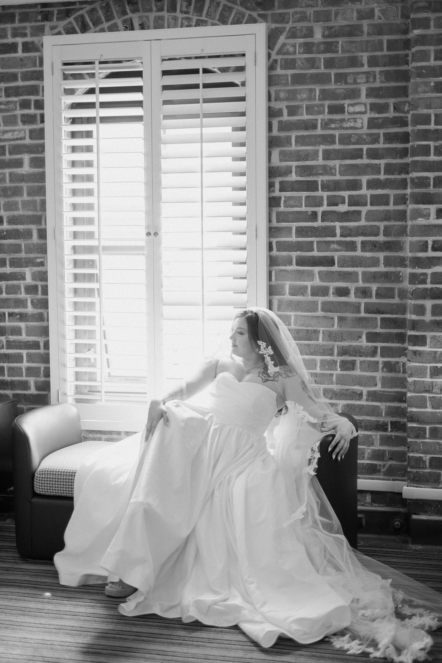 Bride in white gown and veil sits gracefully by shuttered window in black and white bridal portrait.