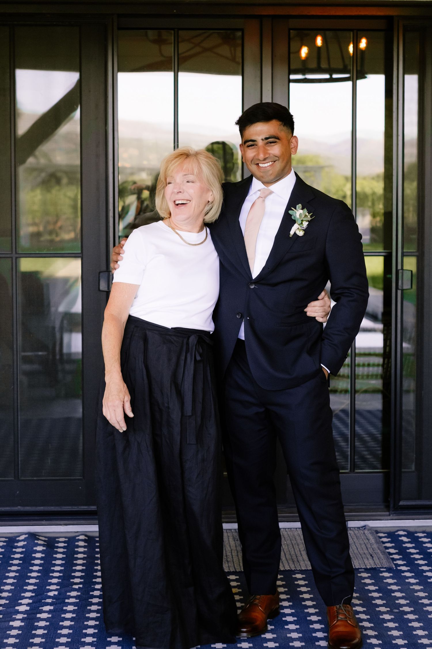 Elderly woman and young groom in navy suit with boutonniere smiling together at outdoor wedding venue.