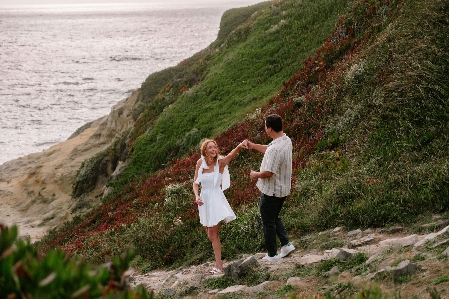Couple dancing on rocky coastal cliffside with green hills and ocean backdrop.