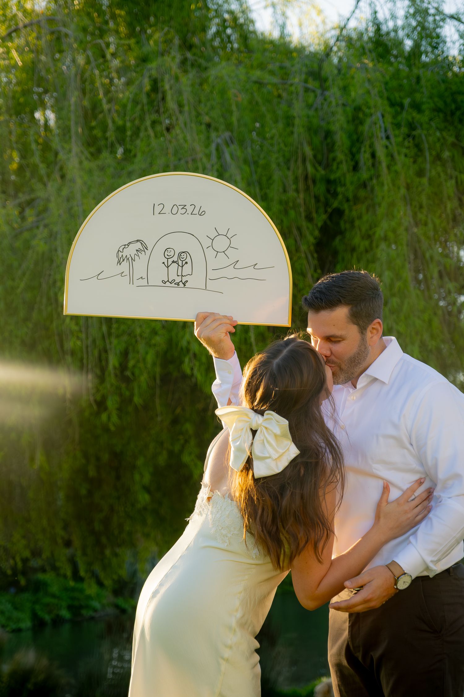 Engaged couple kissing while holding a hand-drawn fan sign showing their wedding date 12.03.26.