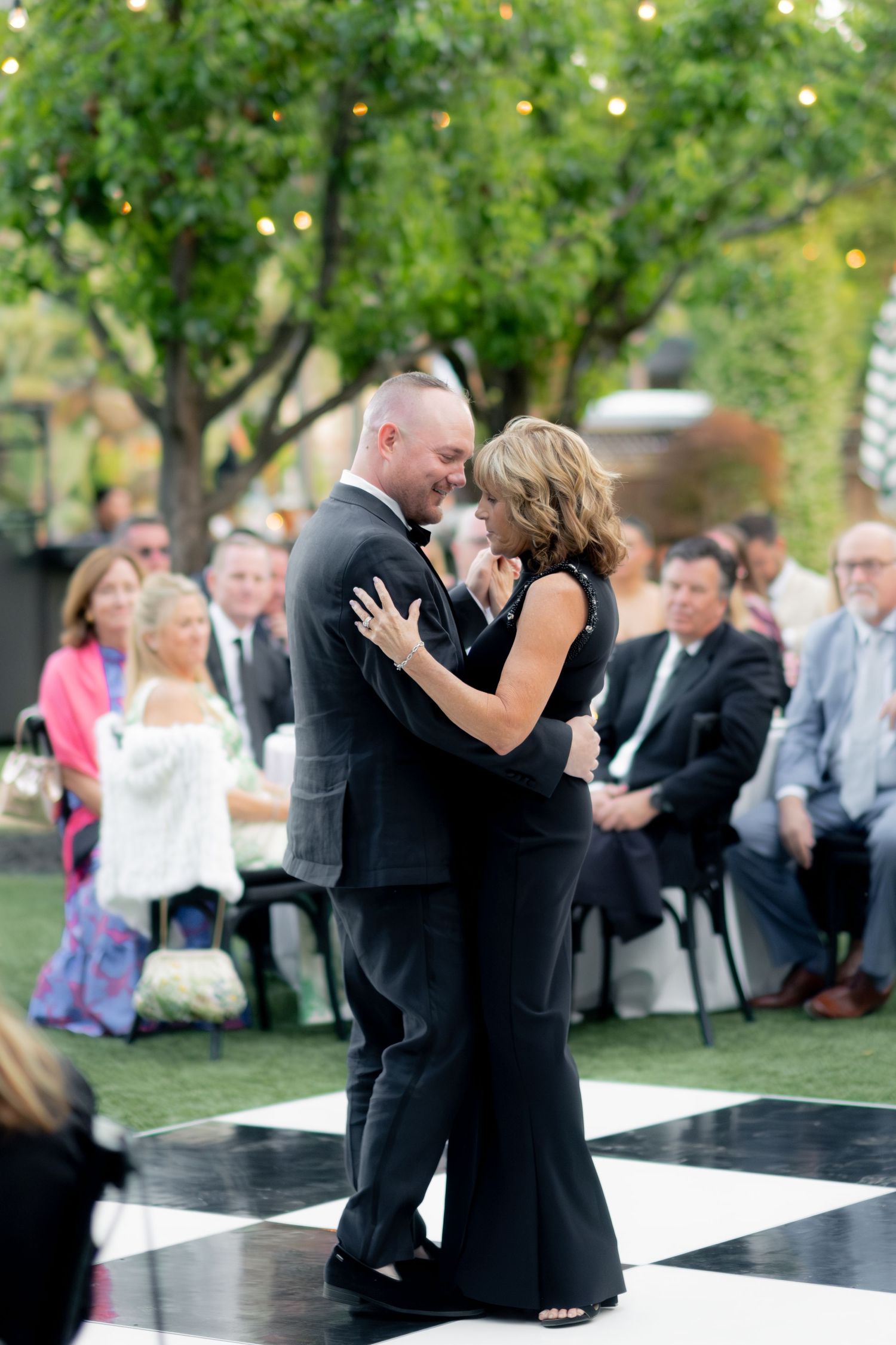 Couple sharing a mother-son dance on a black and white checkered dance floor at an outdoor wedding reception.