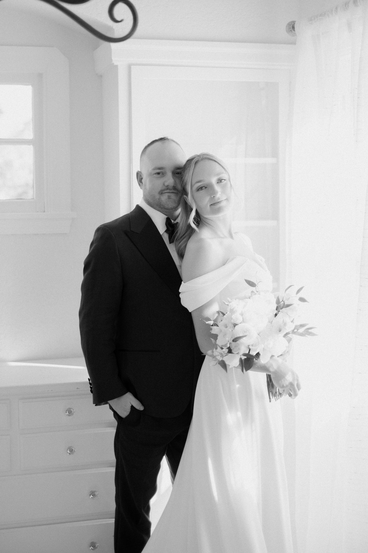 Black and white portrait of bride and groom embracing indoors near bright window.