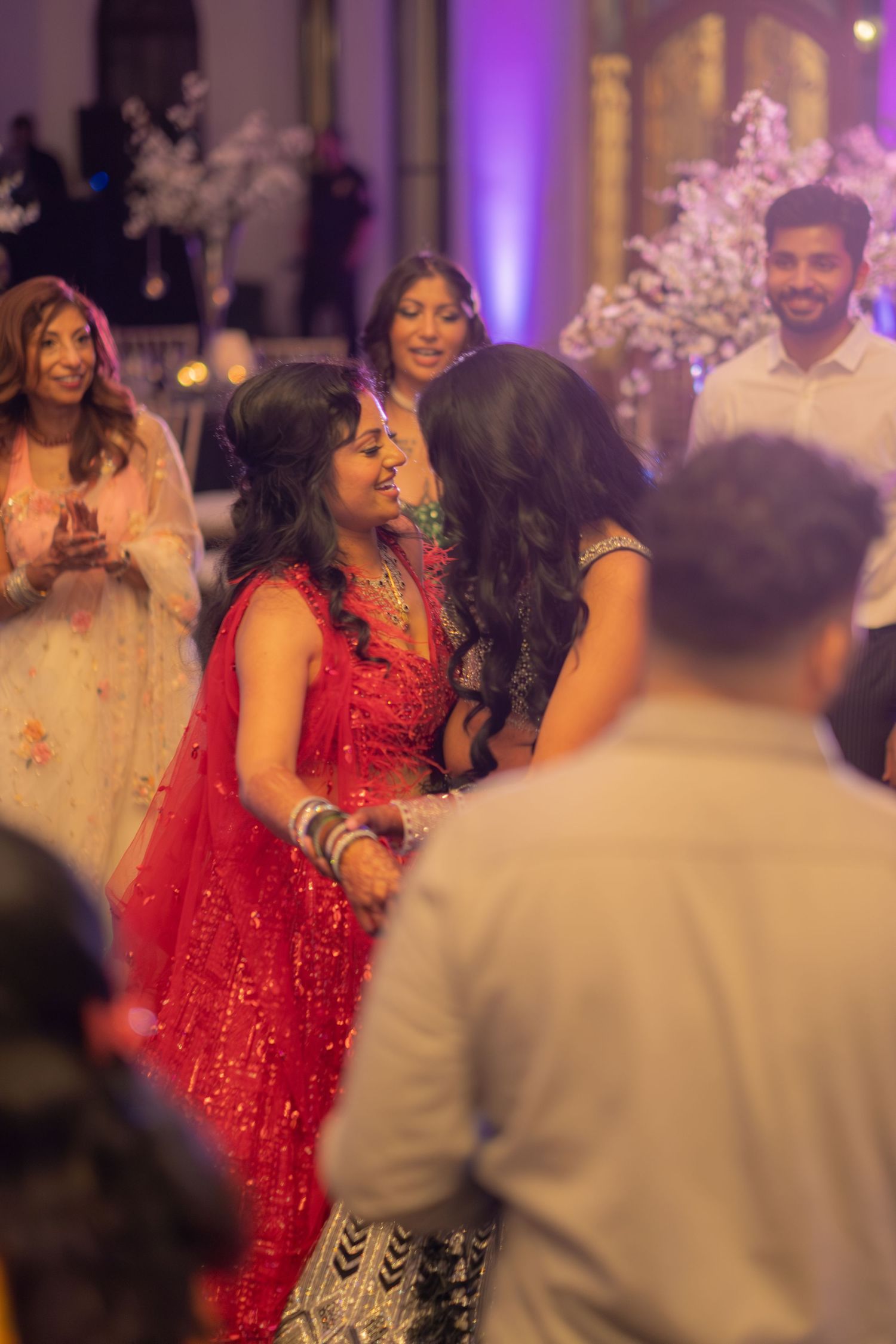 Two women in vibrant Indian lehengas share a joyful moment on the dance floor at a wedding reception.