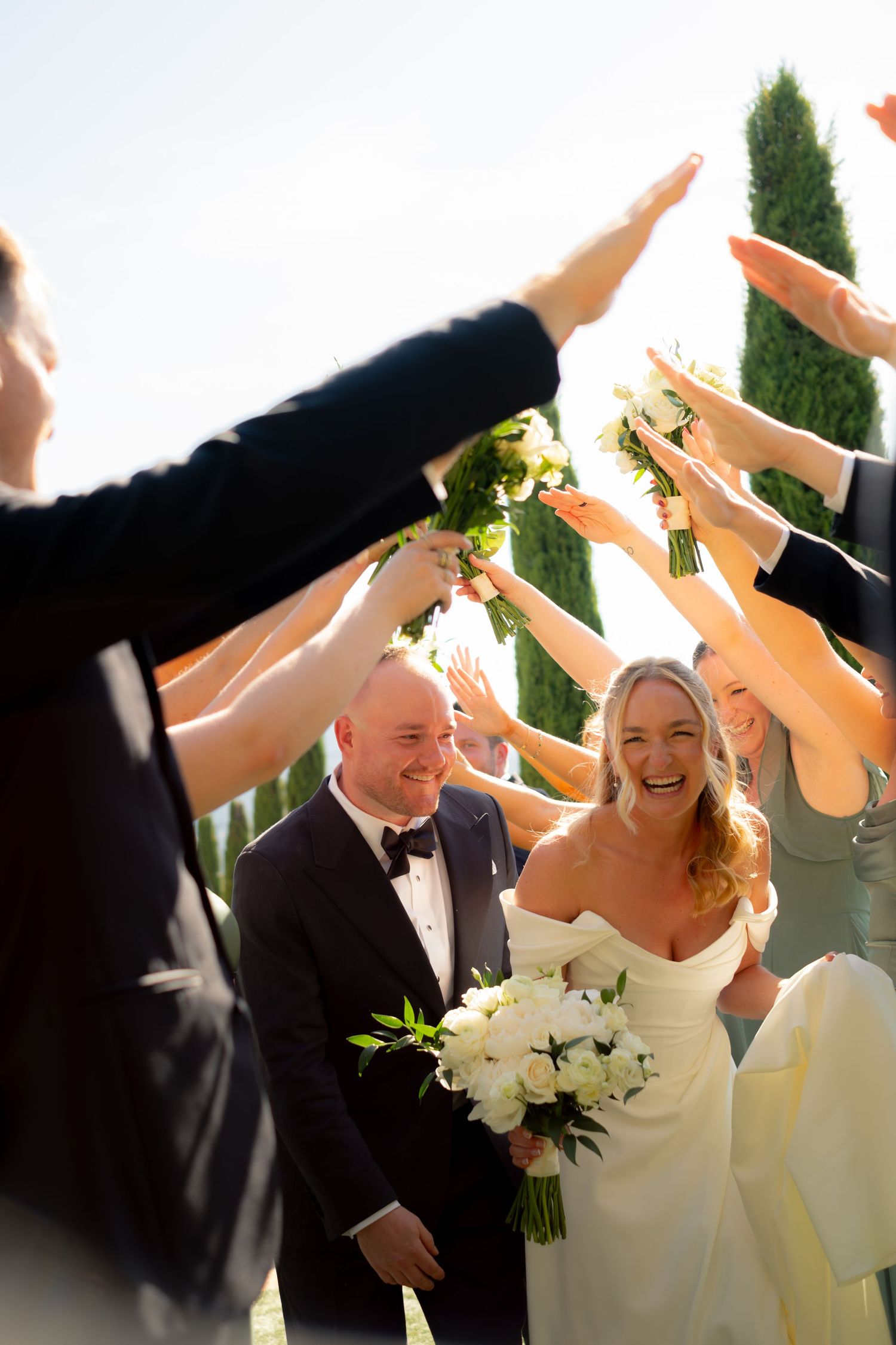 Joyful bride and groom walk through an arch of raised hands formed by their wedding party outdoors.