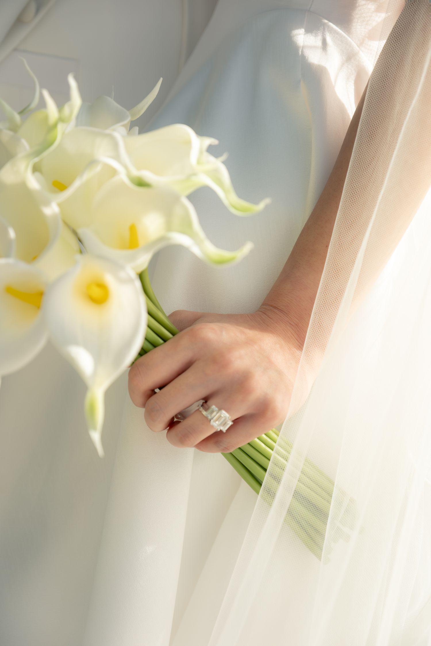 Close-up of bride holding a white calla lily bouquet with a diamond ring visible on her hand.
