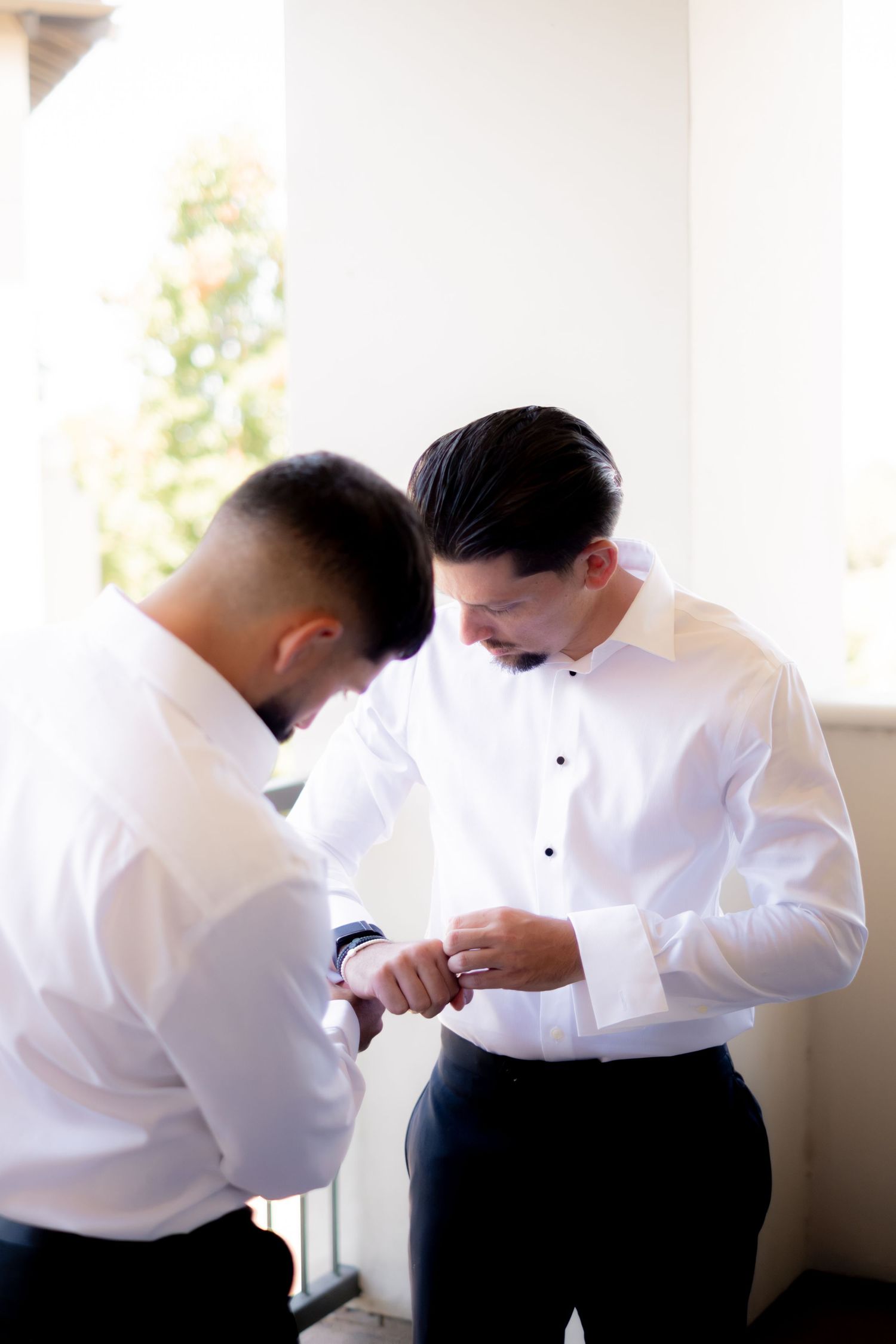 Best man helping groom fasten cufflinks while getting ready in white dress shirts before wedding ceremony.
