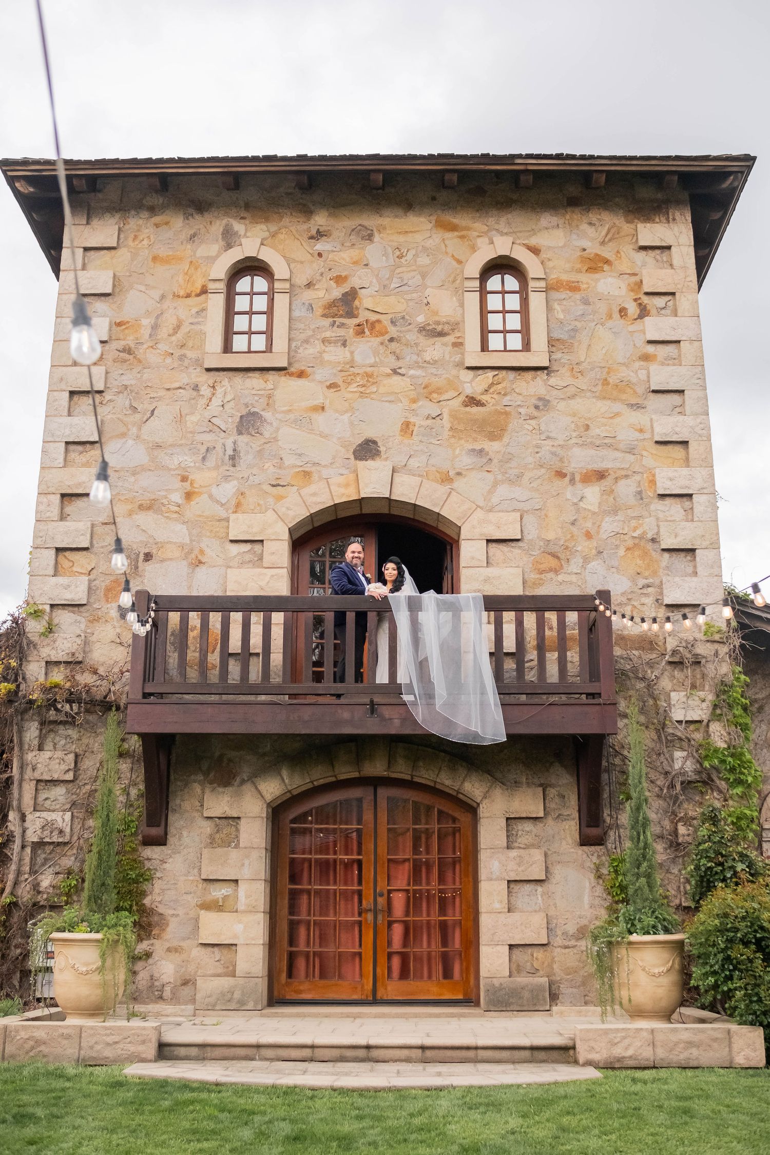 Bride and groom posing on balcony of rustic stone tower building at winery wedding venue.