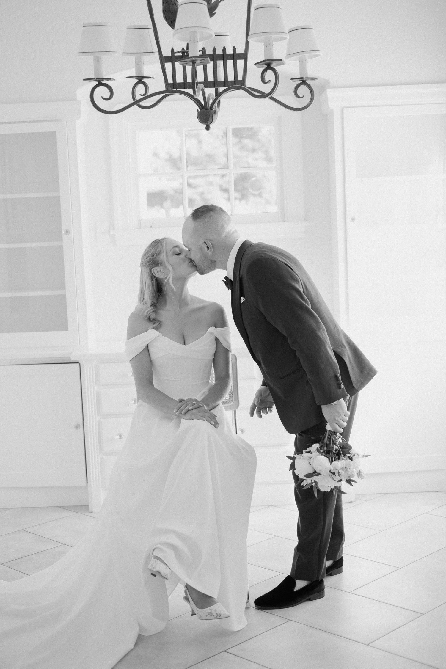 Black and white romantic photo of groom kissing bride in off-shoulder gown beneath chandelier in bright white room.