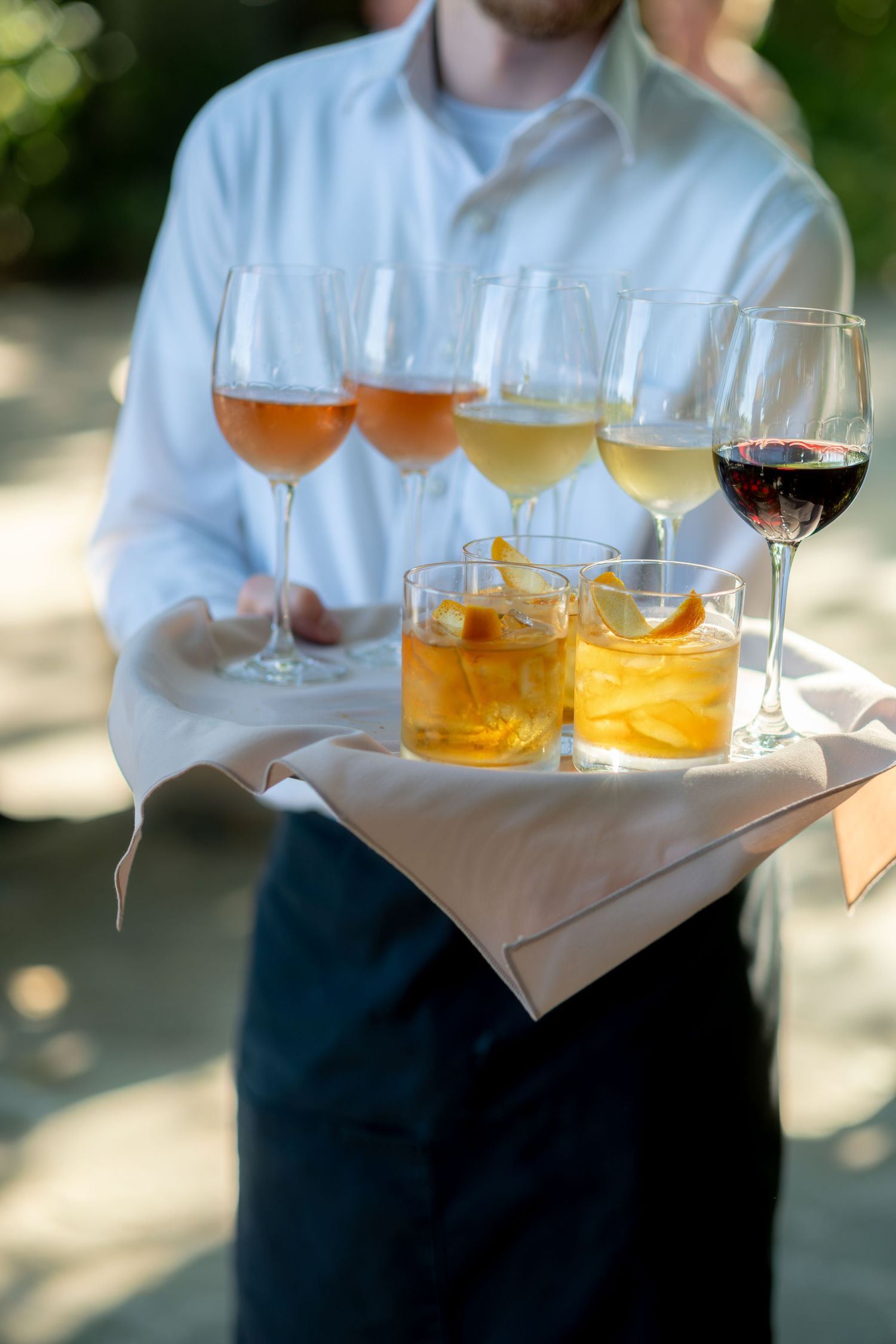Waiter carries tray of cocktails including old fashioneds and wine glasses at outdoor wedding reception.