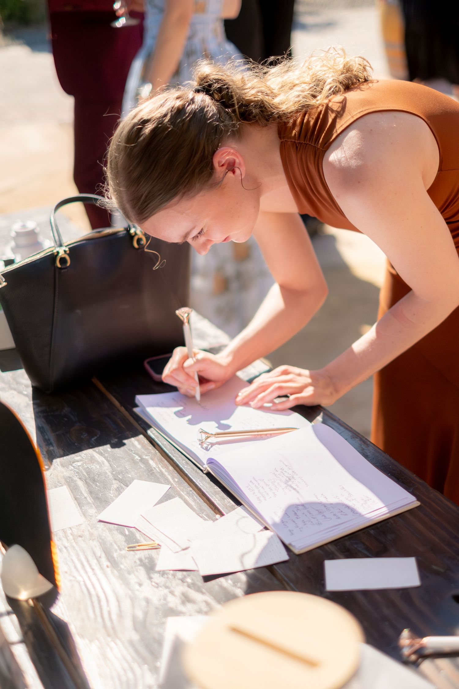 A woman in an orange dress signs a guest book at a sunny outdoor wedding reception table.