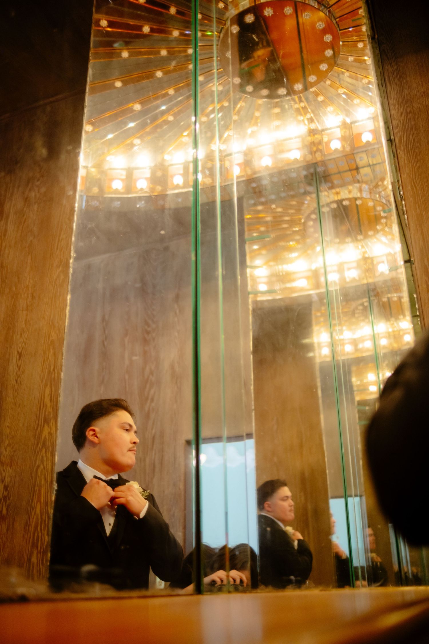 Groom adjusting bow tie in ornate mirrored elevator with golden chandelier ceiling reflection.