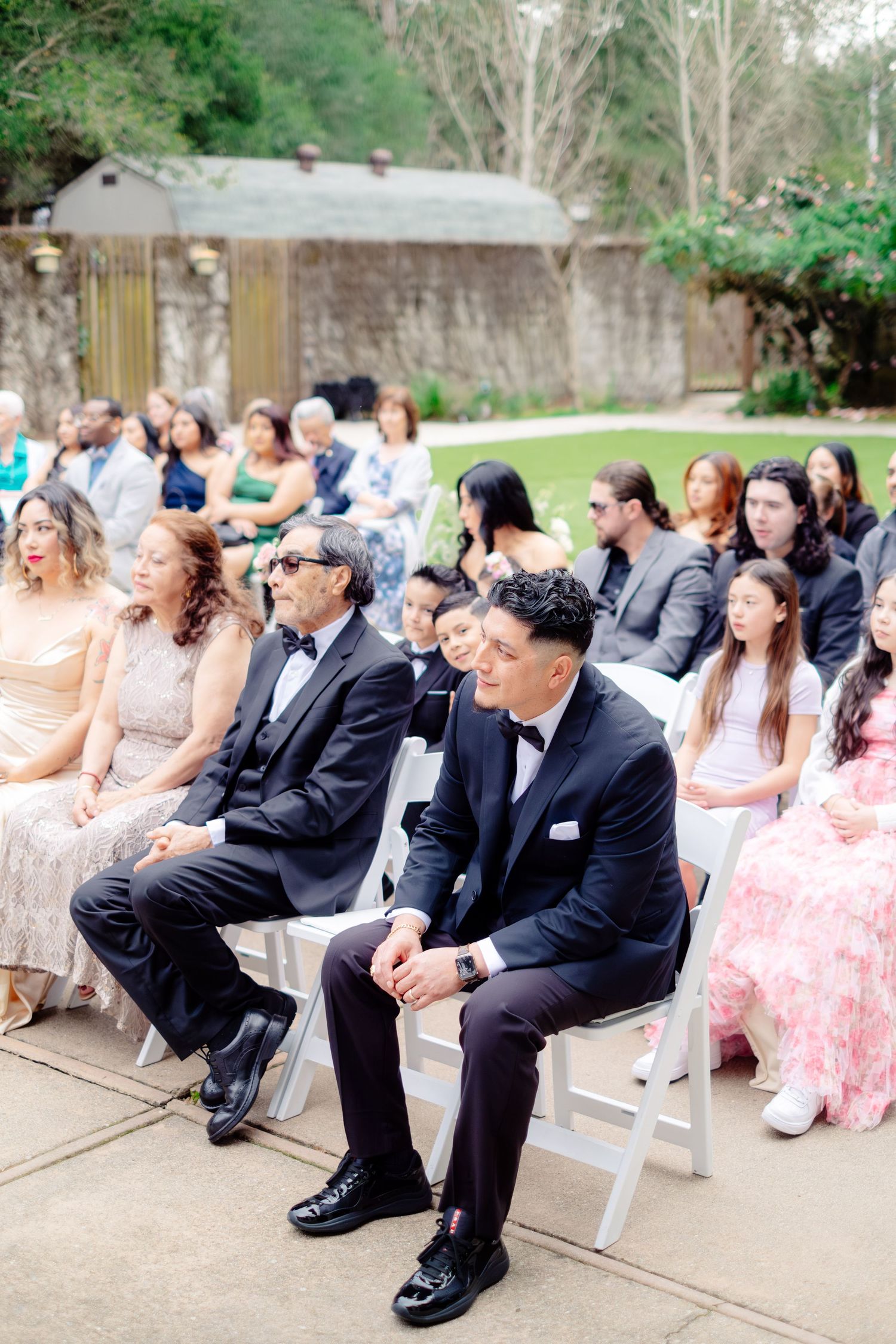 Wedding guests in formal attire seated outdoors watching ceremony in garden venue.