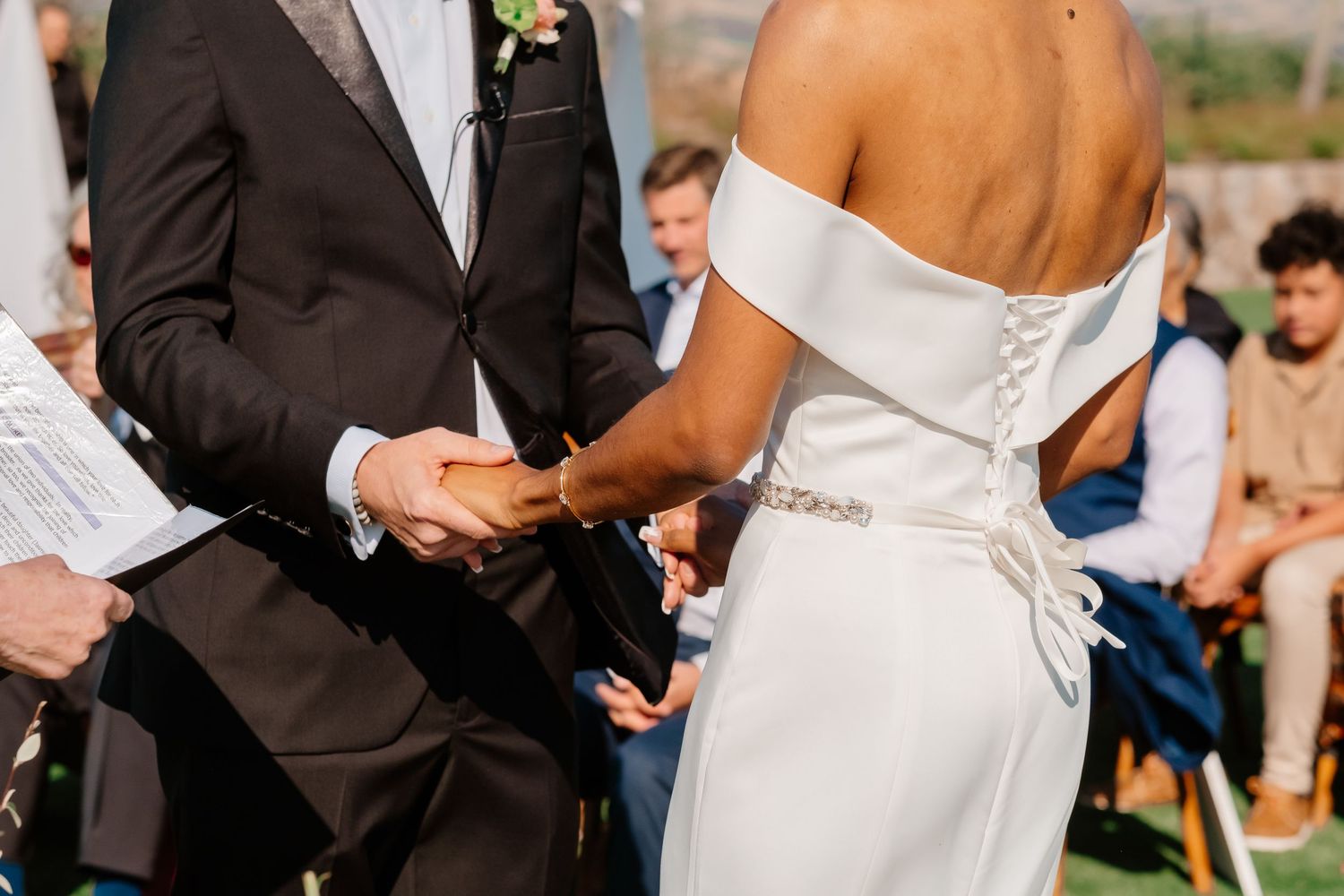 Bride and groom holding hands during outdoor wedding ceremony, bride wearing elegant off-shoulder white gown with lace back.