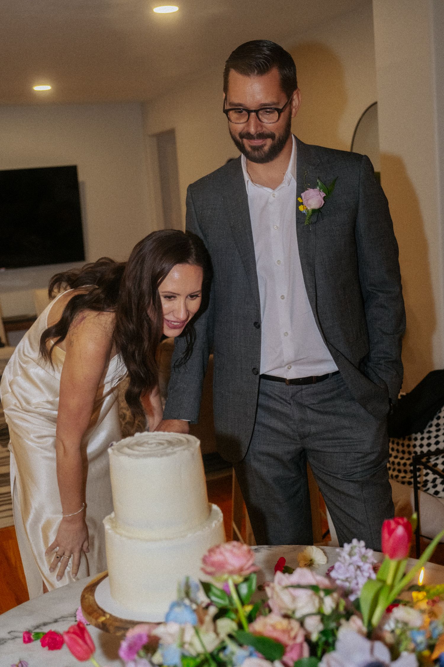 Bride and groom smiling while cutting a two-tier white wedding cake surrounded by colorful floral arrangements.