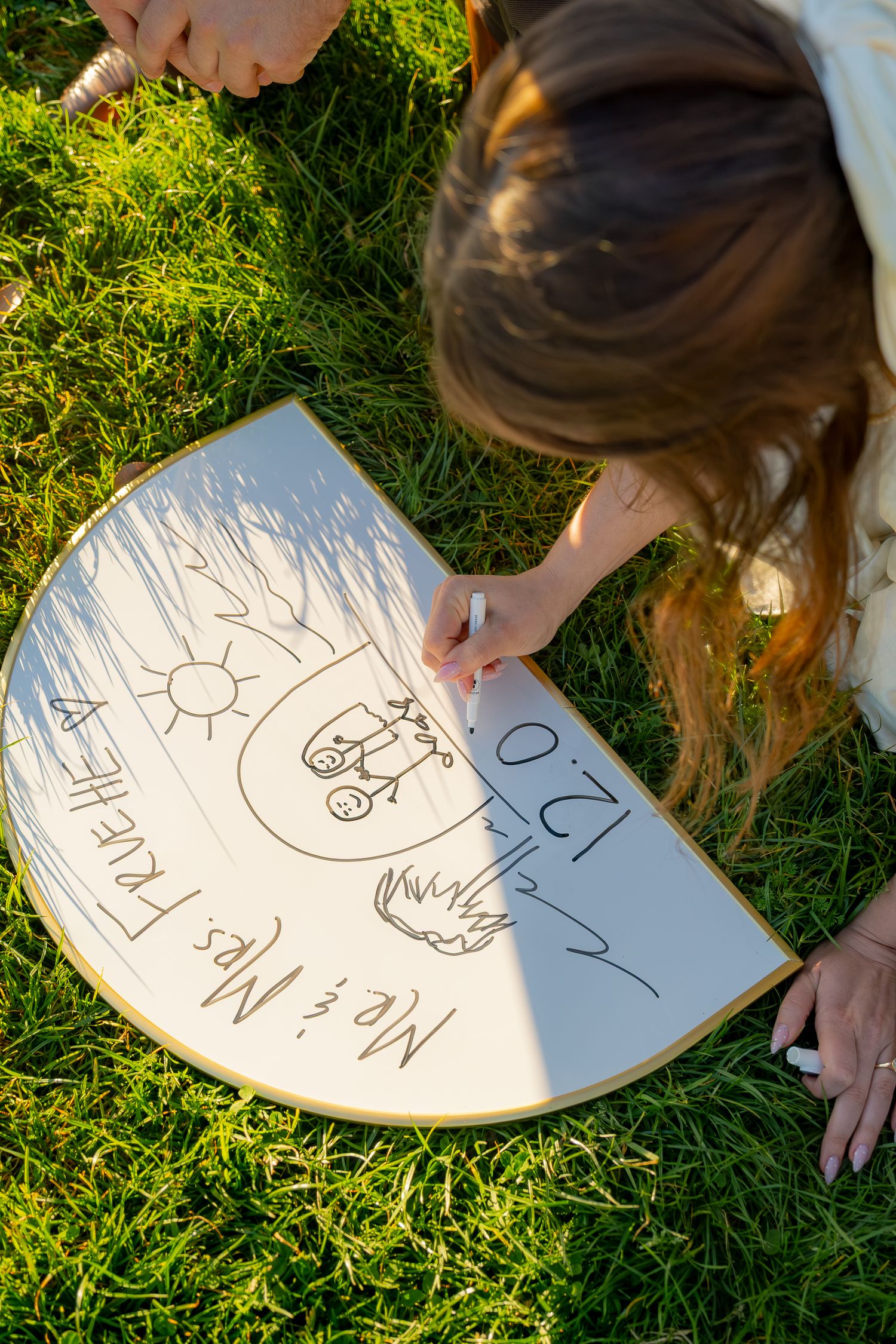 Bride-to-be writing wedding date on a decorative fan-shaped sign while lying on green grass.