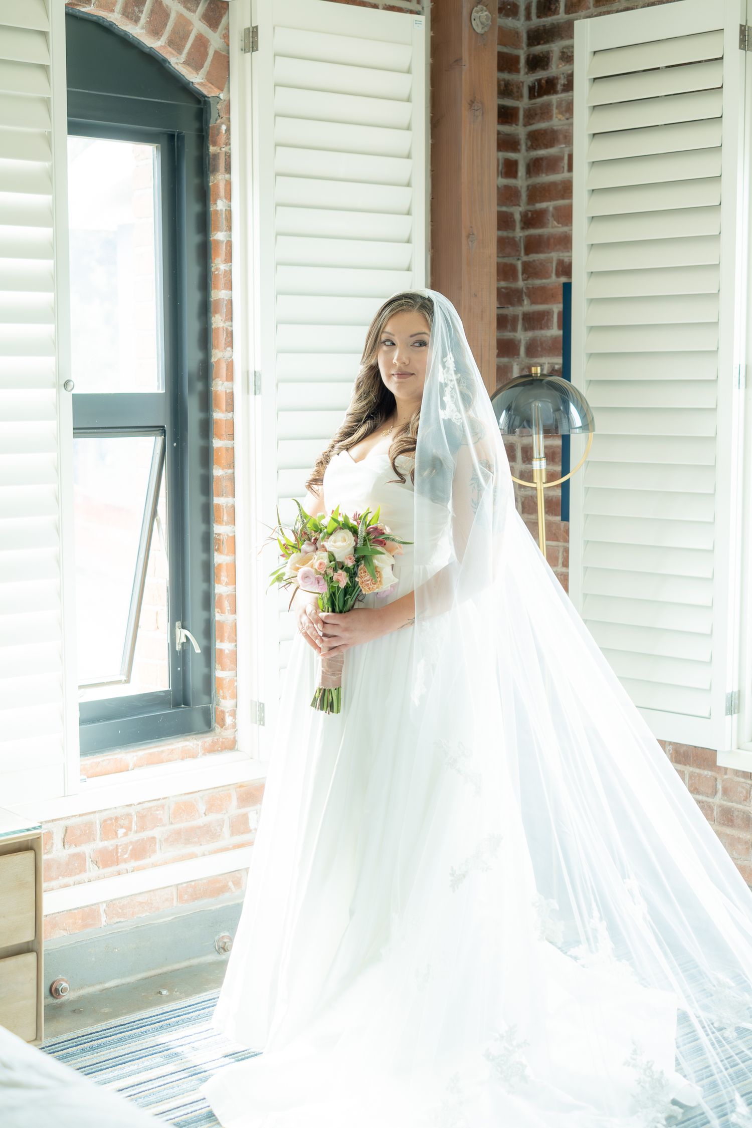 Bride in white gown and veil holding pink bouquet standing by window in brick venue.