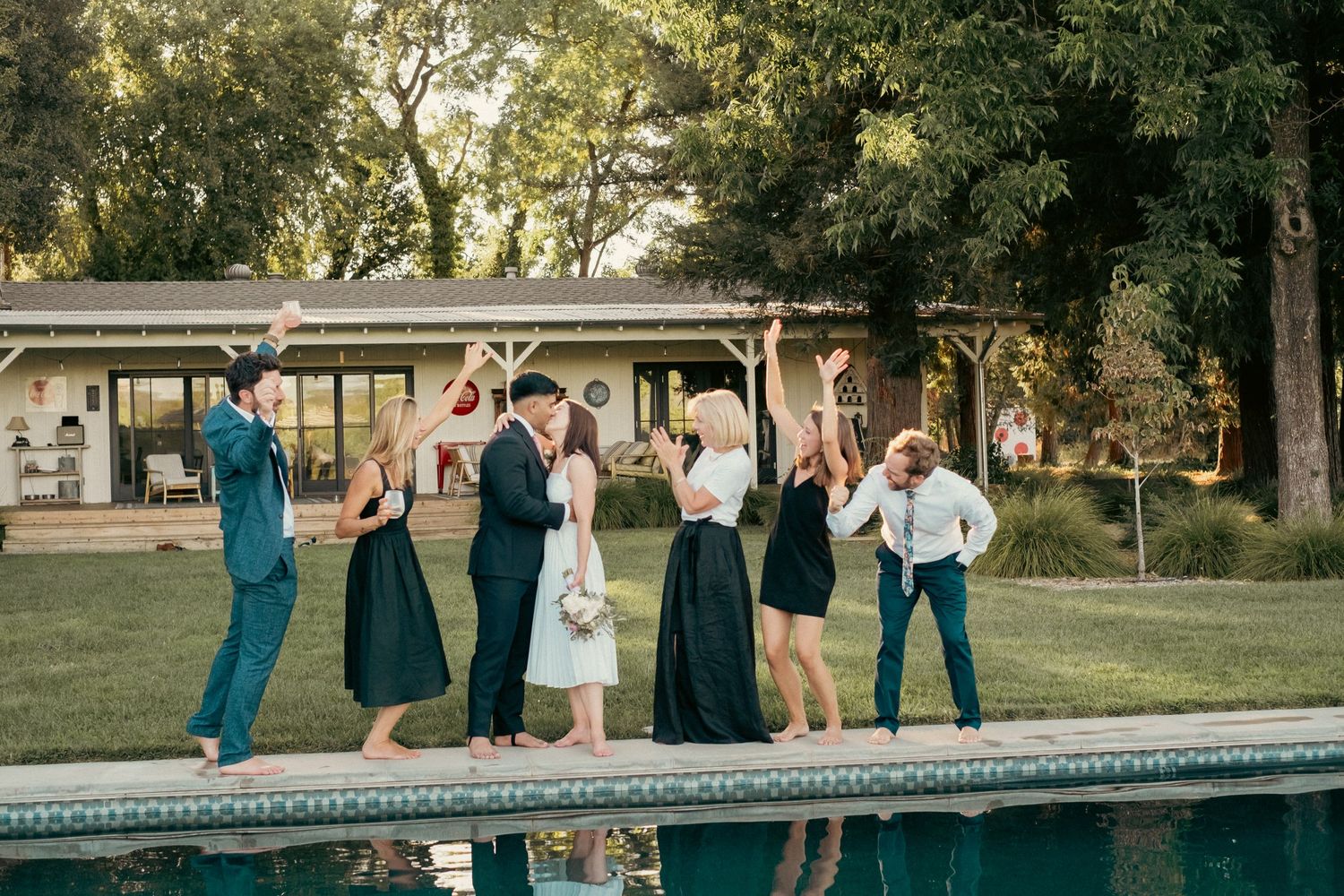 Wedding party celebrating poolside in elegant attire, dancing outdoors near a modern house surrounded by lush trees.