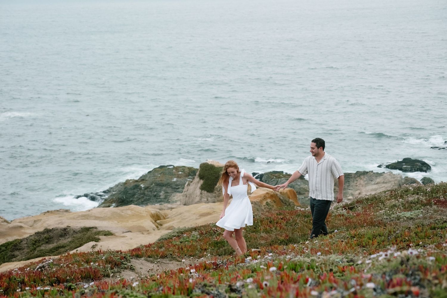 Couple holding hands on a rocky coastal cliff with ocean waves and wildflowers in the background.