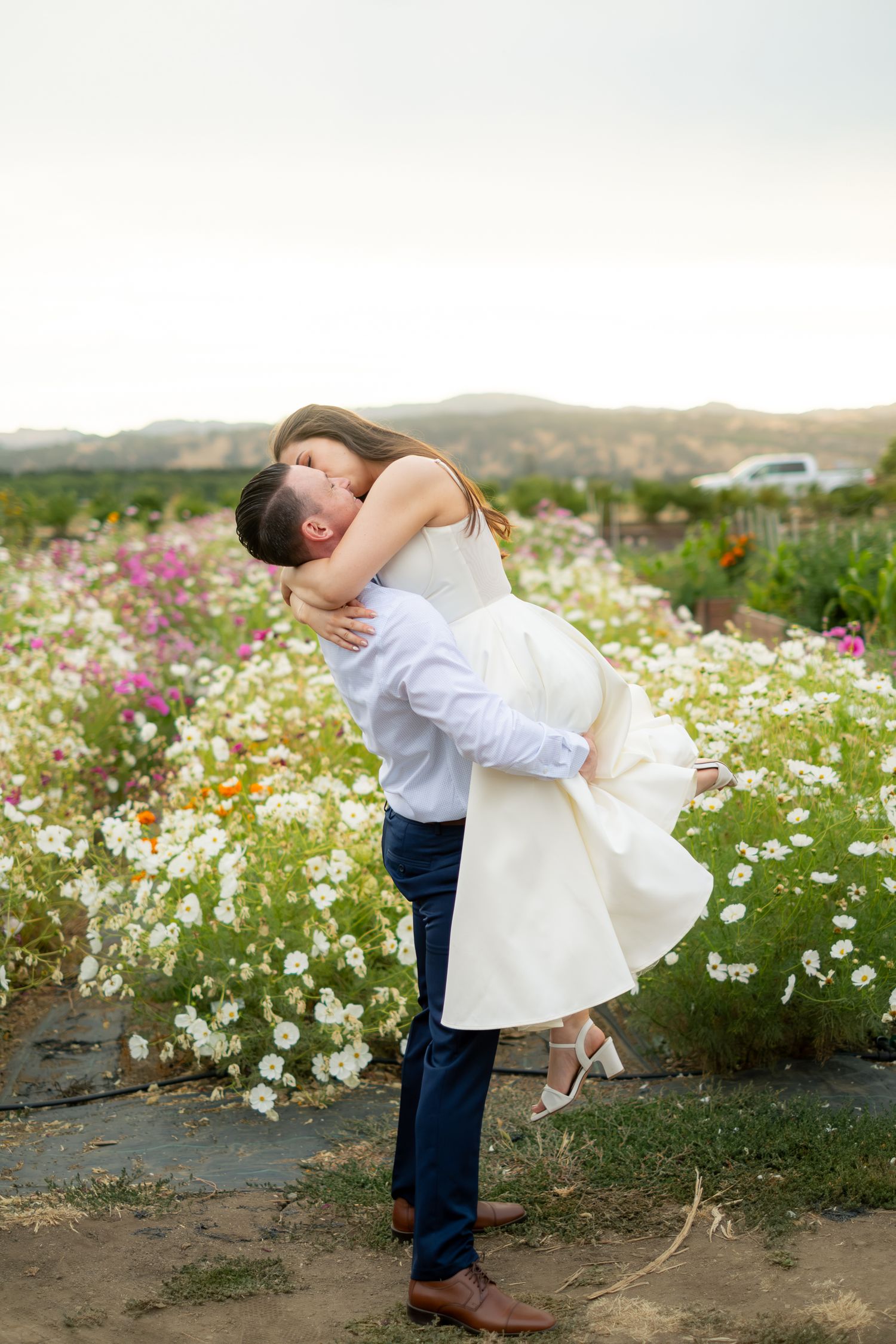 Groom lifting bride in white dress for a kiss in a colorful flower field at sunset.