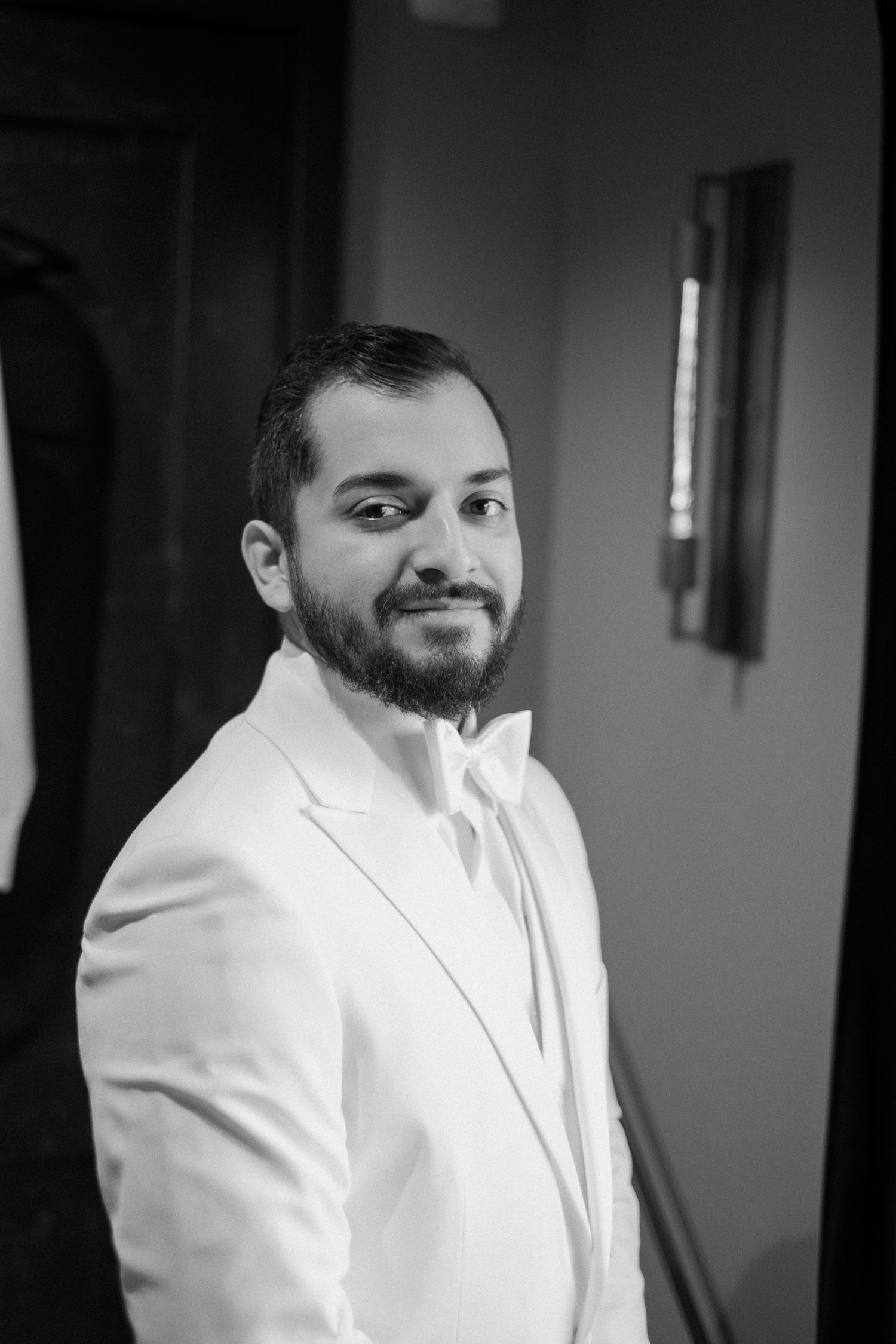 Black and white portrait of a bearded groom in a white suit jacket smiling over his shoulder.