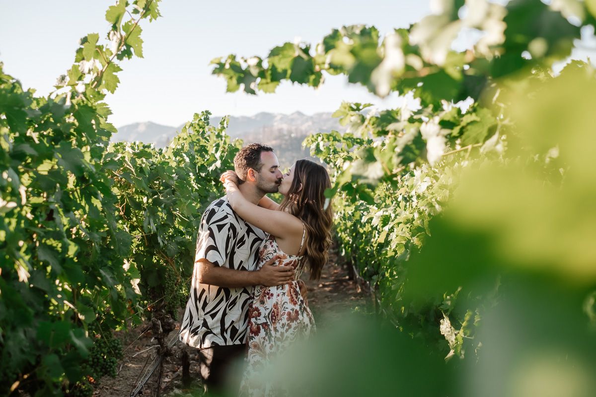 Couple sharing a romantic kiss among lush green vineyard rows with mountains in the background.