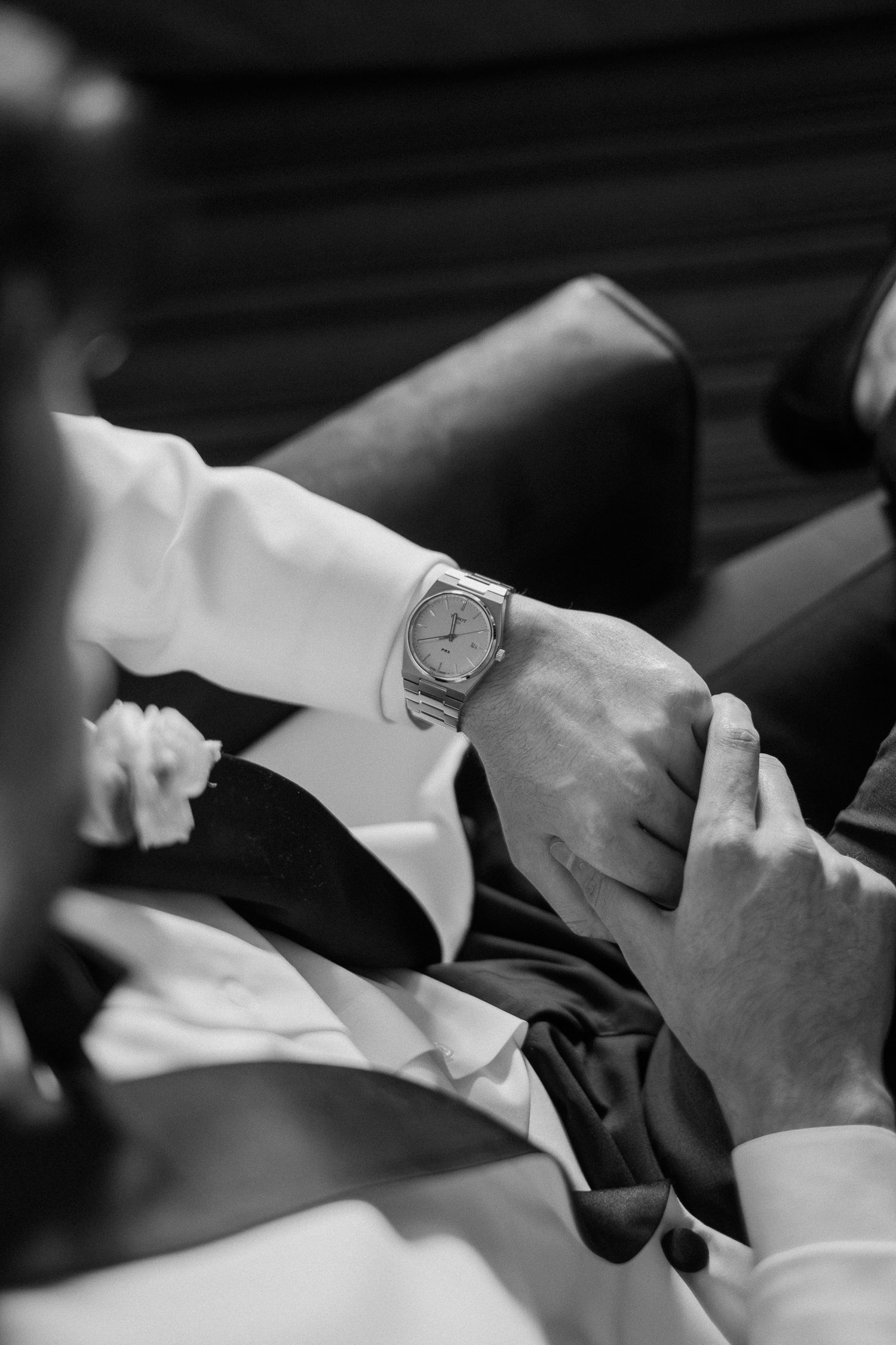 Black and white photo of a groom adjusting his luxury wristwatch, wearing a suit with boutonniere.
