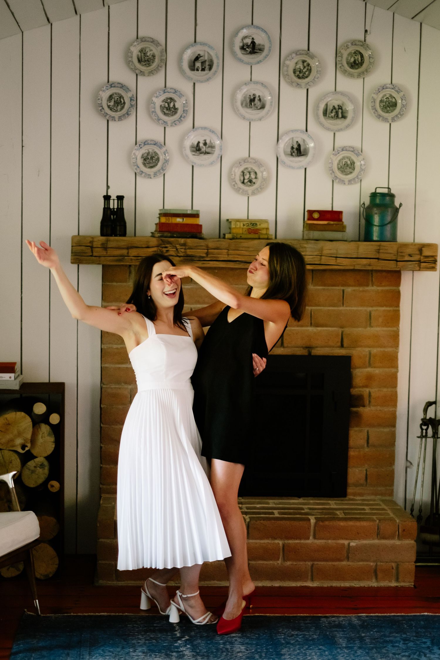 Two women dancing and laughing in front of a brick fireplace, one in a white dress and one in a black dress.
