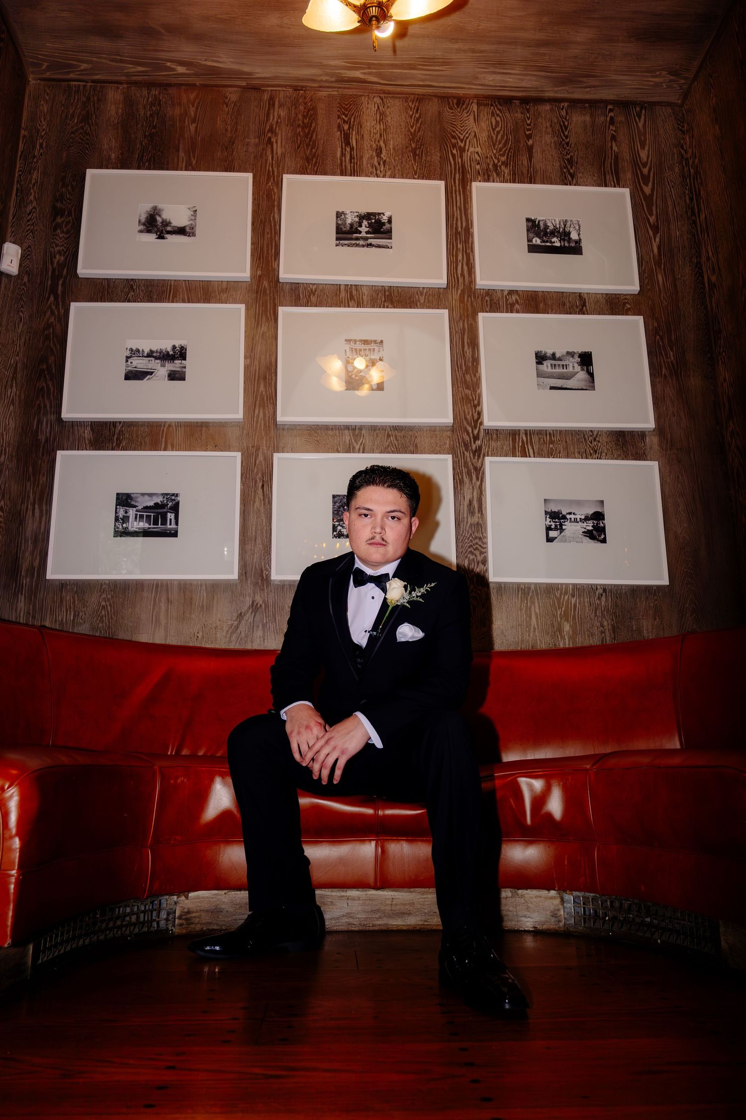 Groom in black tuxedo sits on red leather sofa beneath framed black and white photos.
