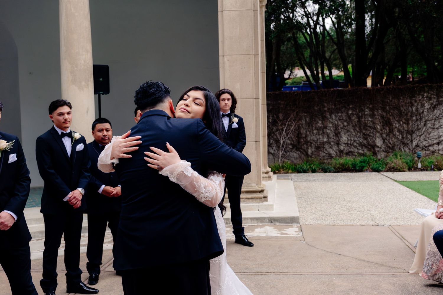 Bride and groom share an emotional hug during outdoor wedding ceremony with groomsmen in black tuxedos standing nearby.