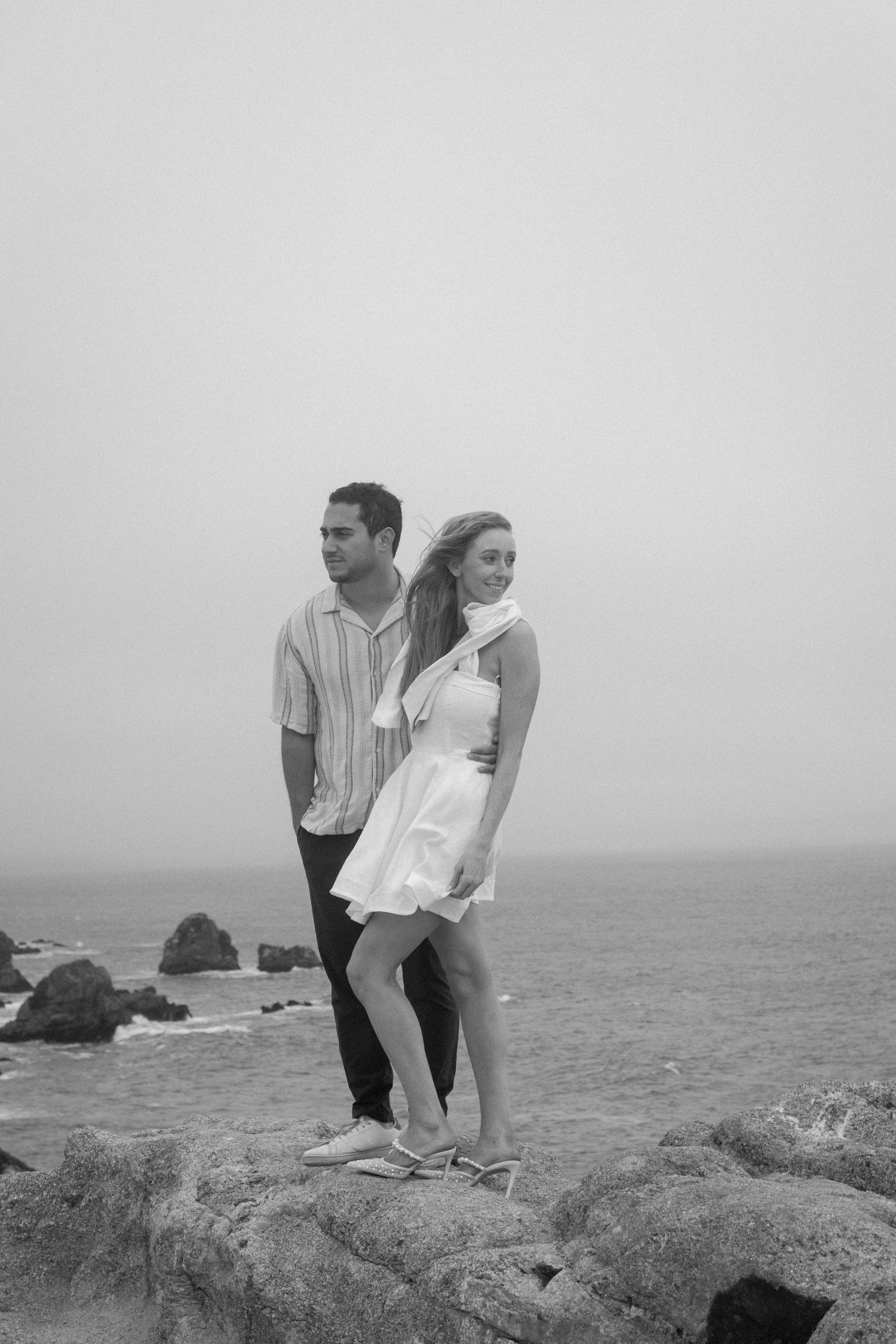 Black and white photo of a couple standing on rocky coastal cliffs with ocean waves in the background.