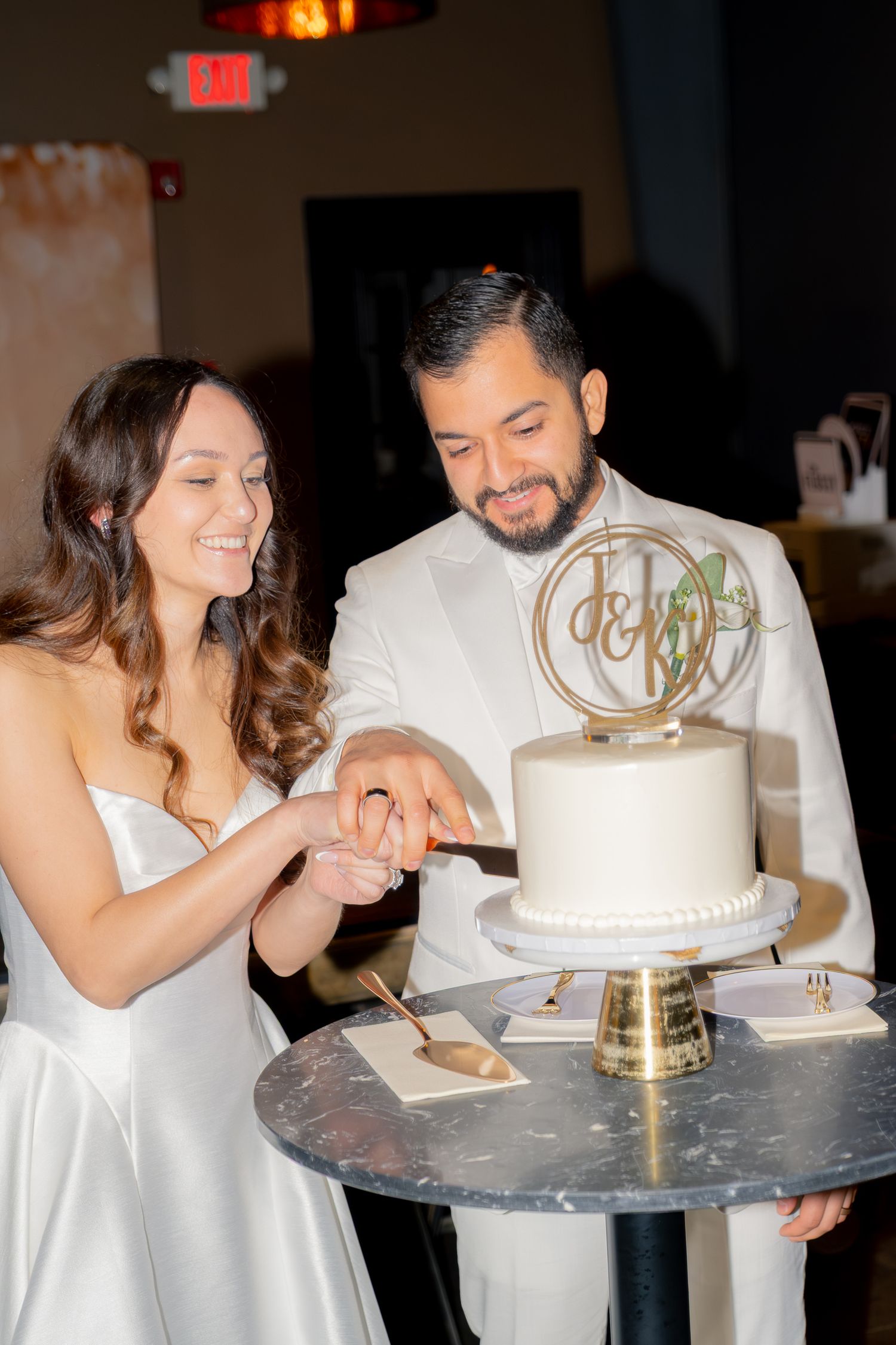 Bride and groom cutting elegant white single-tier wedding cake with gold monogram topper at reception.
