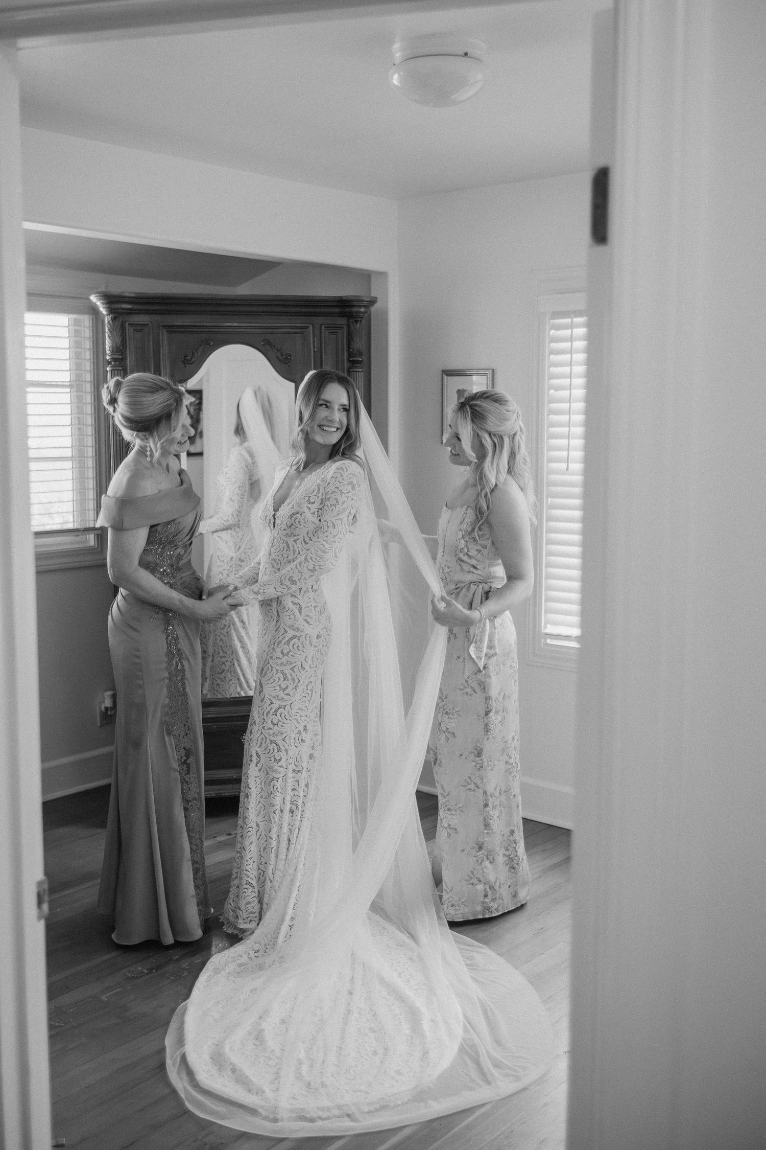 Black and white photo of bride in lace gown with long veil assisted by two bridesmaids in elegant dresses.