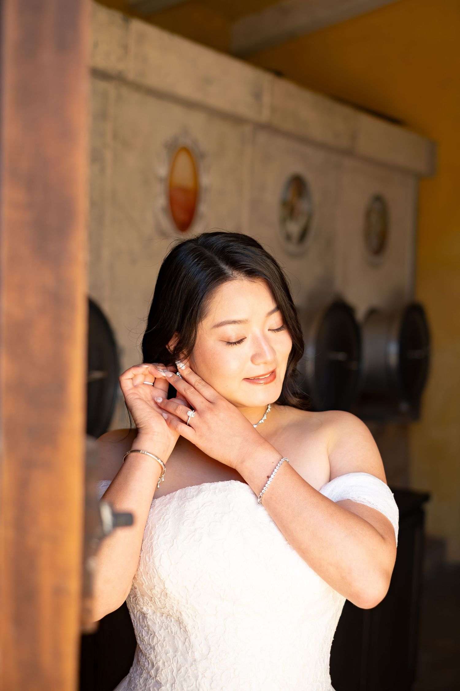 Bride in off-shoulder white gown adjusts earring with eyes closed in warm golden light.