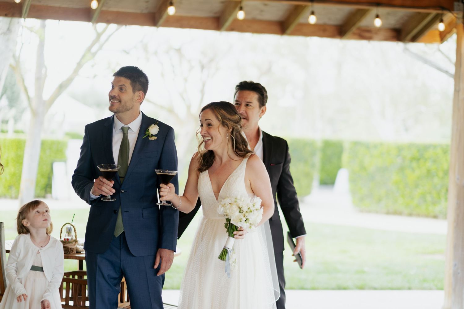 Bride in white gown walks with two groomsmen in navy suits under string lights at outdoor wedding venue.
