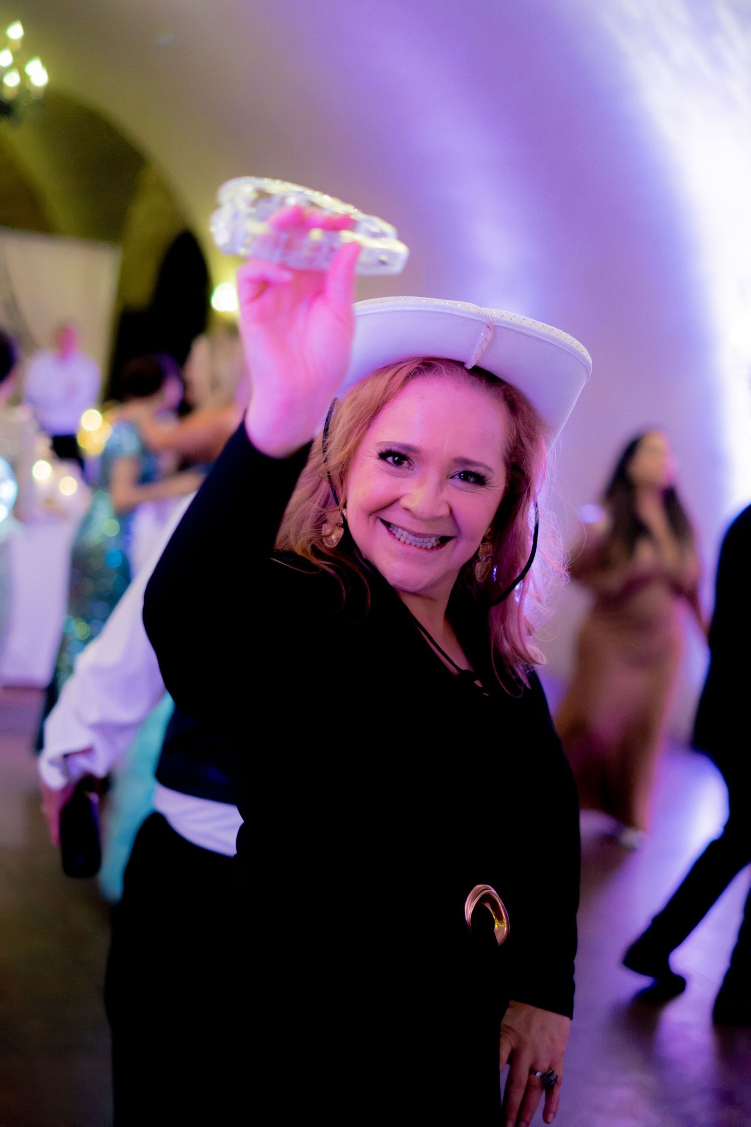 Joyful woman in black outfit holds white cowboy hat and tambourine on wedding dance floor.