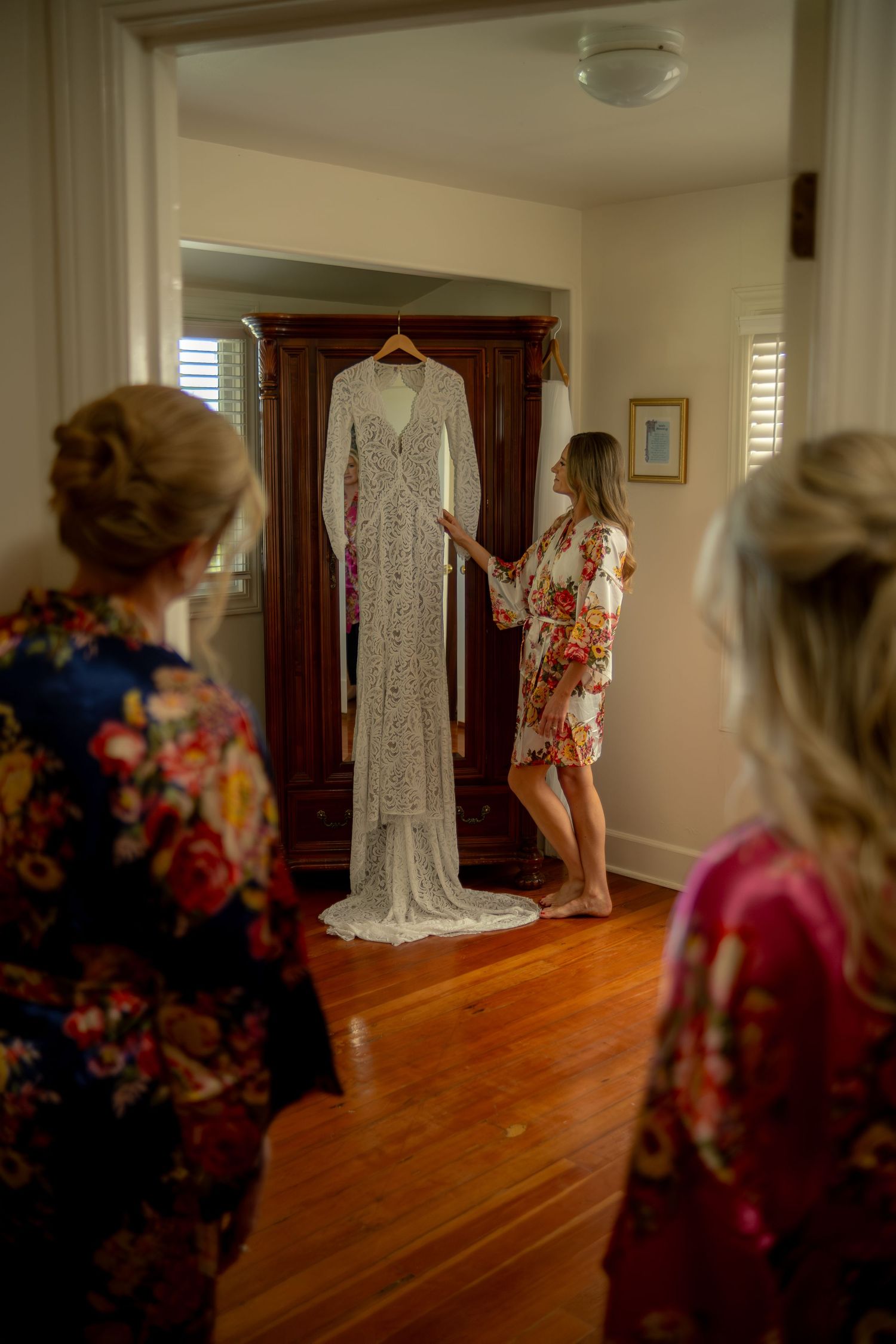 Bride in floral robe admires lace wedding gown on armoire while bridesmaids look on.