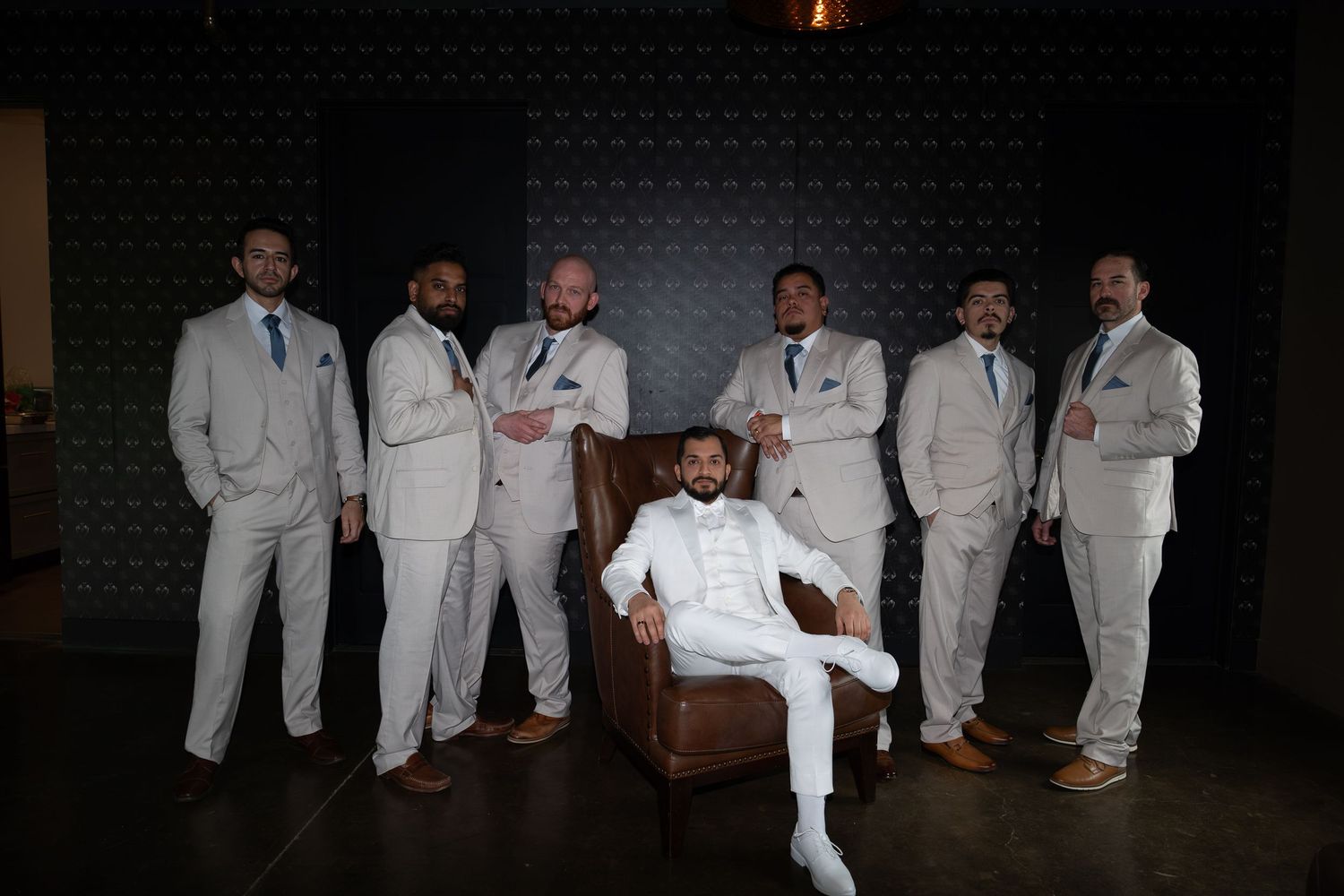 Groom in white suit seated in leather chair surrounded by groomsmen in matching light gray suits posing for wedding photo.