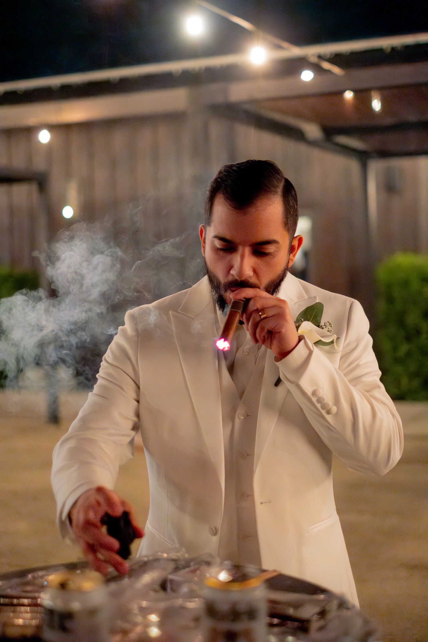 Dapper groom in white suit lighting a cigar at night under string lights at wedding.