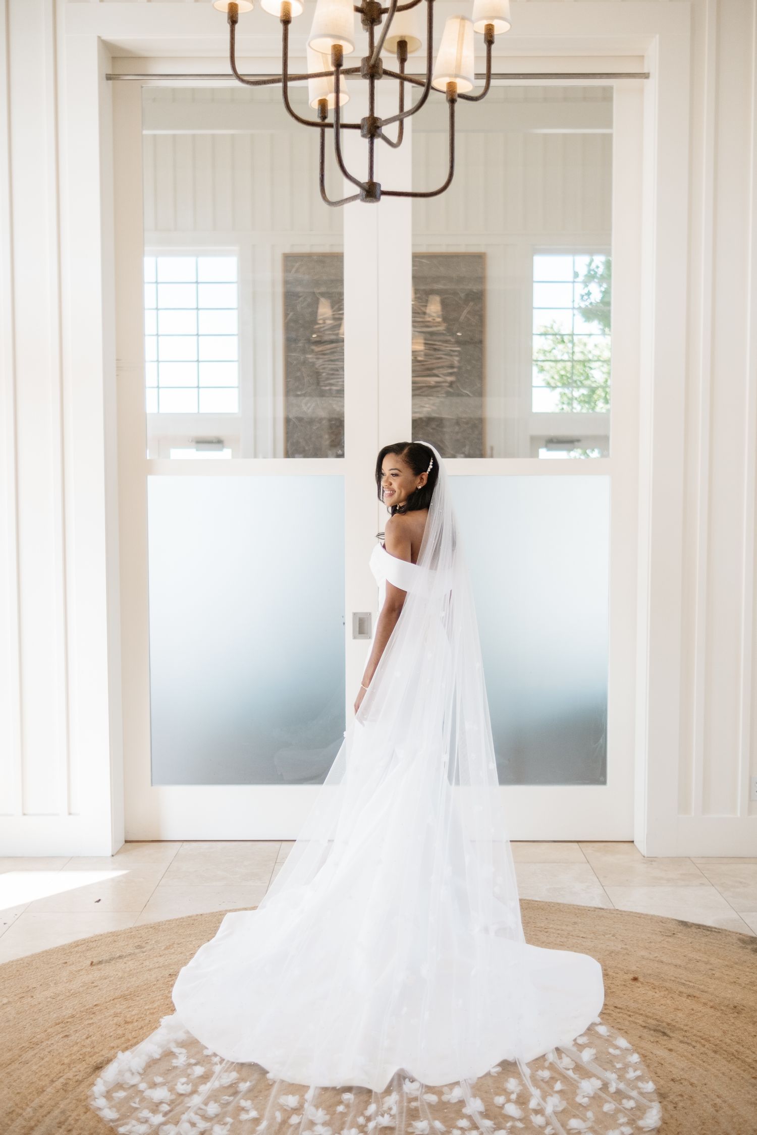 Elegant bride in an off-shoulder lace wedding gown with a long floral veil stands in a bright white room.