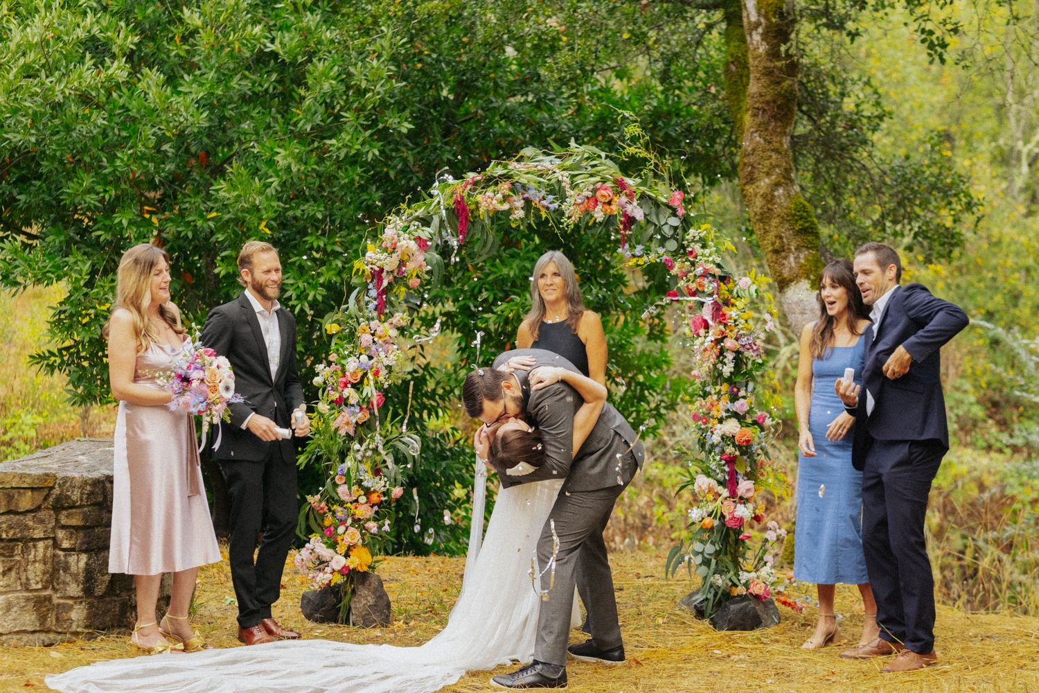 Couple shares first kiss under floral arch at outdoor wedding ceremony with guests watching.