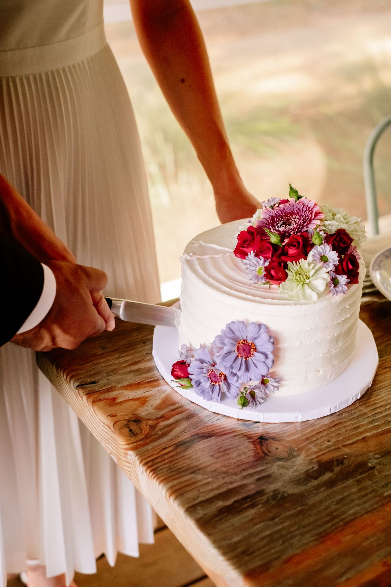 Bride and groom cutting a white wedding cake decorated with colorful fresh flowers on a rustic wooden table.