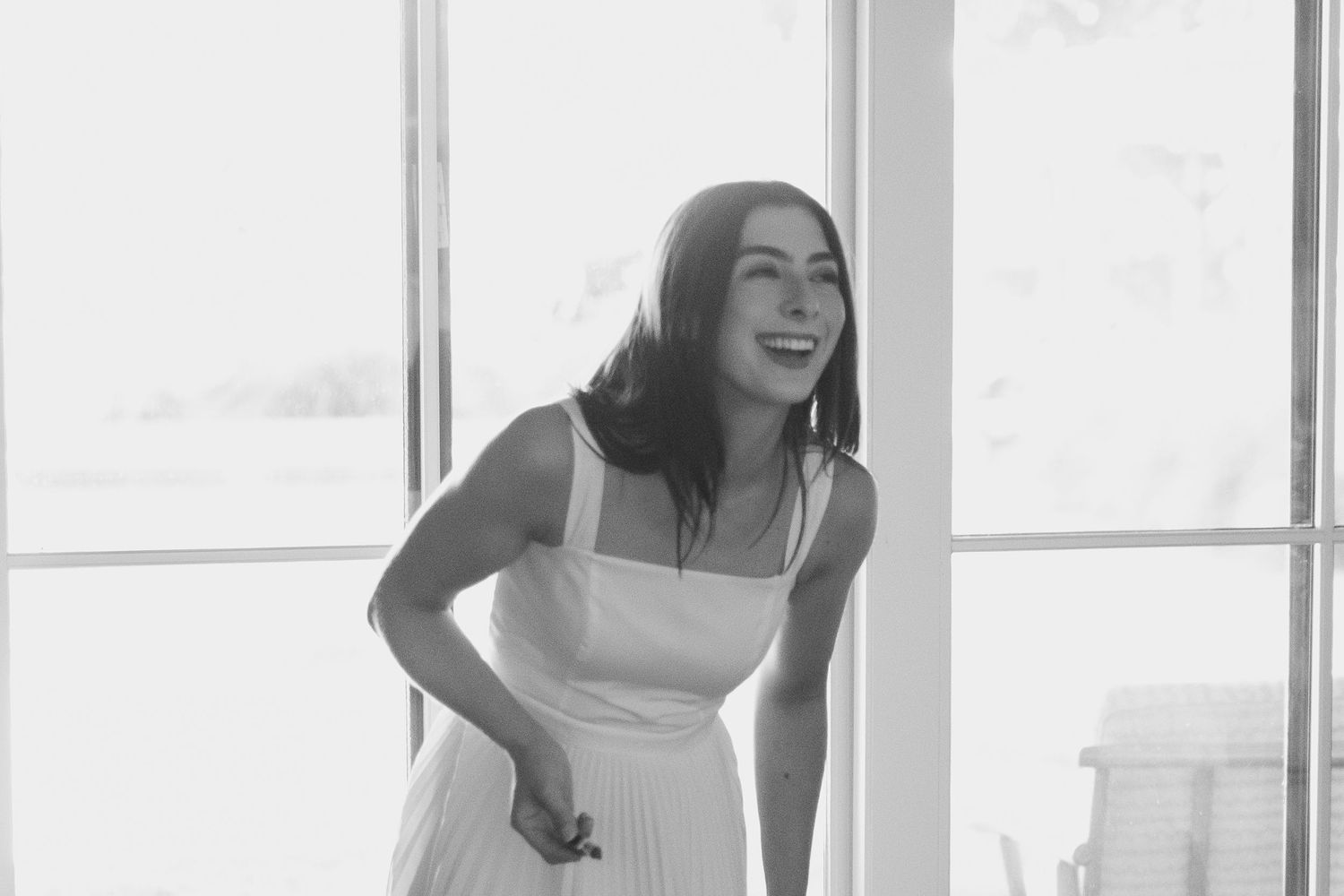 A joyful woman in a white dress laughing by a bright window in a black and white photo.