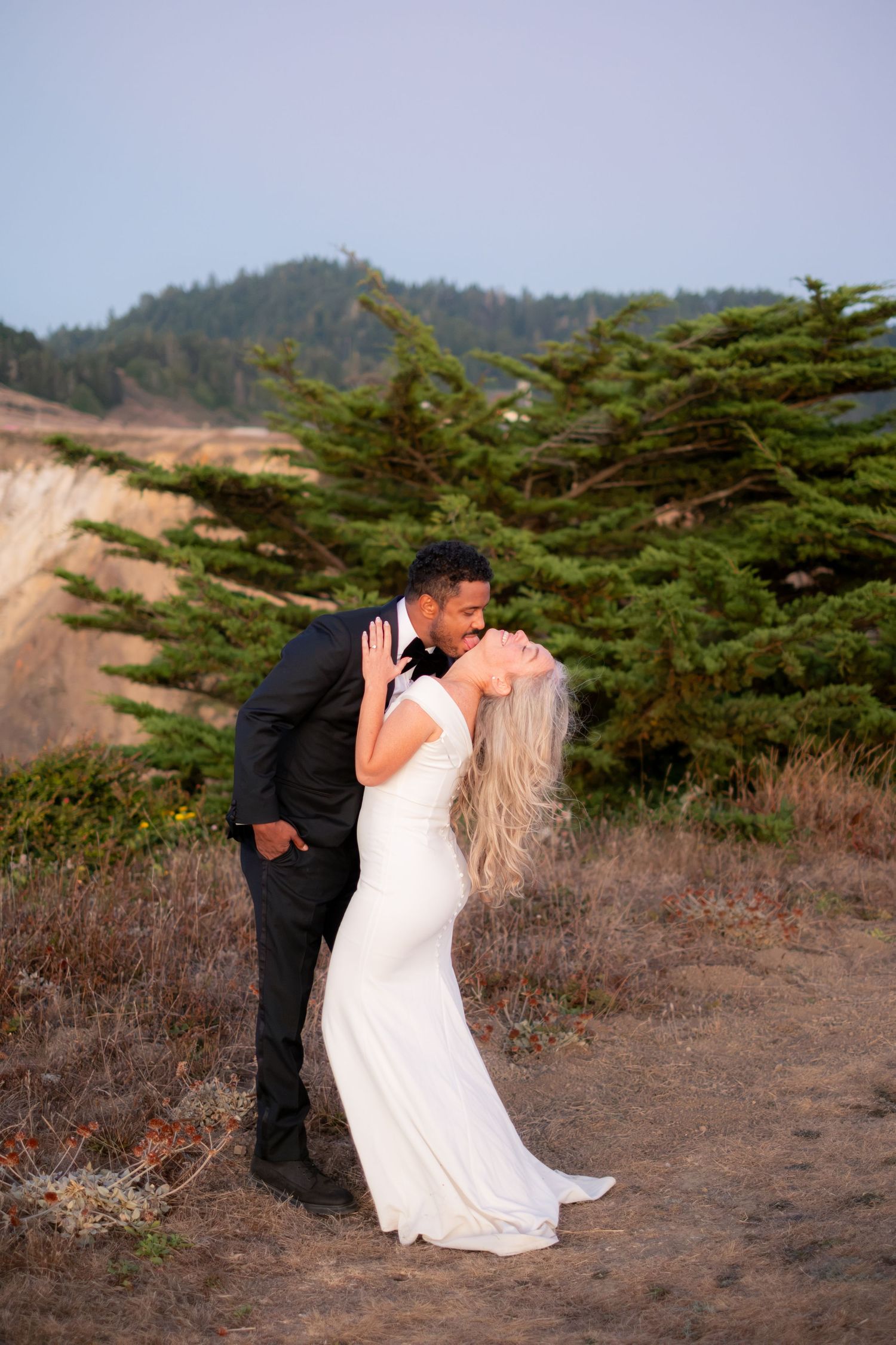 Interracial wedding couple sharing a romantic kiss in a scenic coastal landscape at dusk.