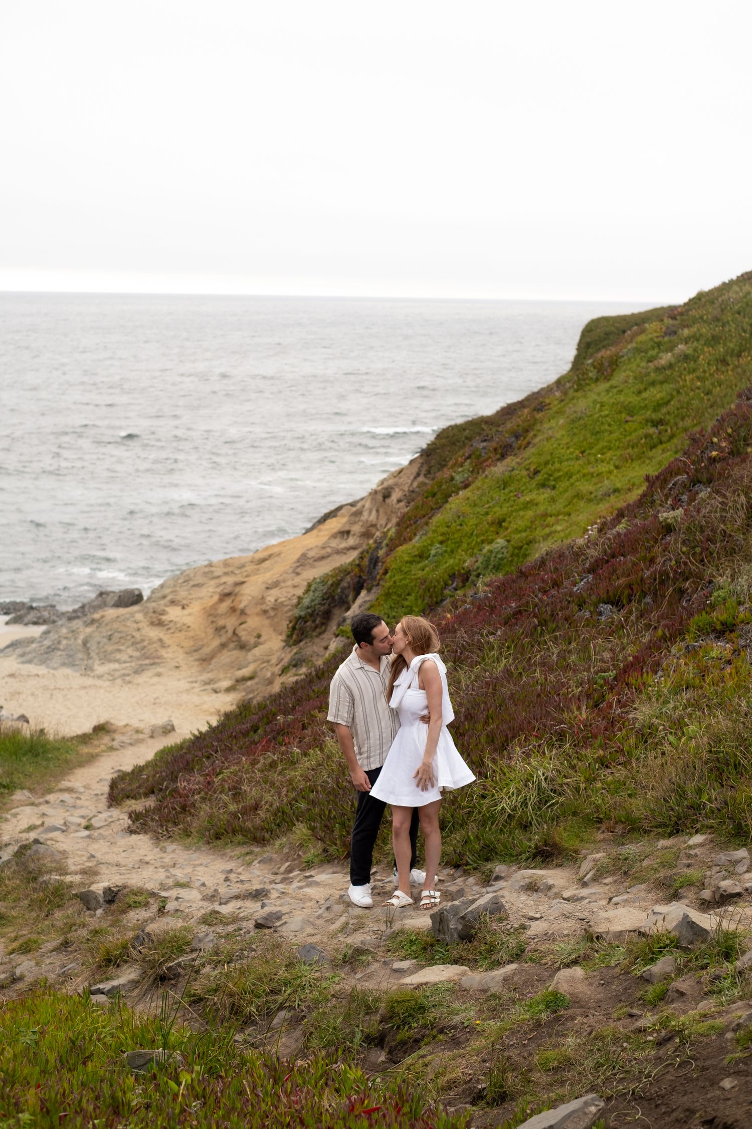 Couple sharing a kiss on a scenic coastal trail with green cliffs and ocean views behind them.