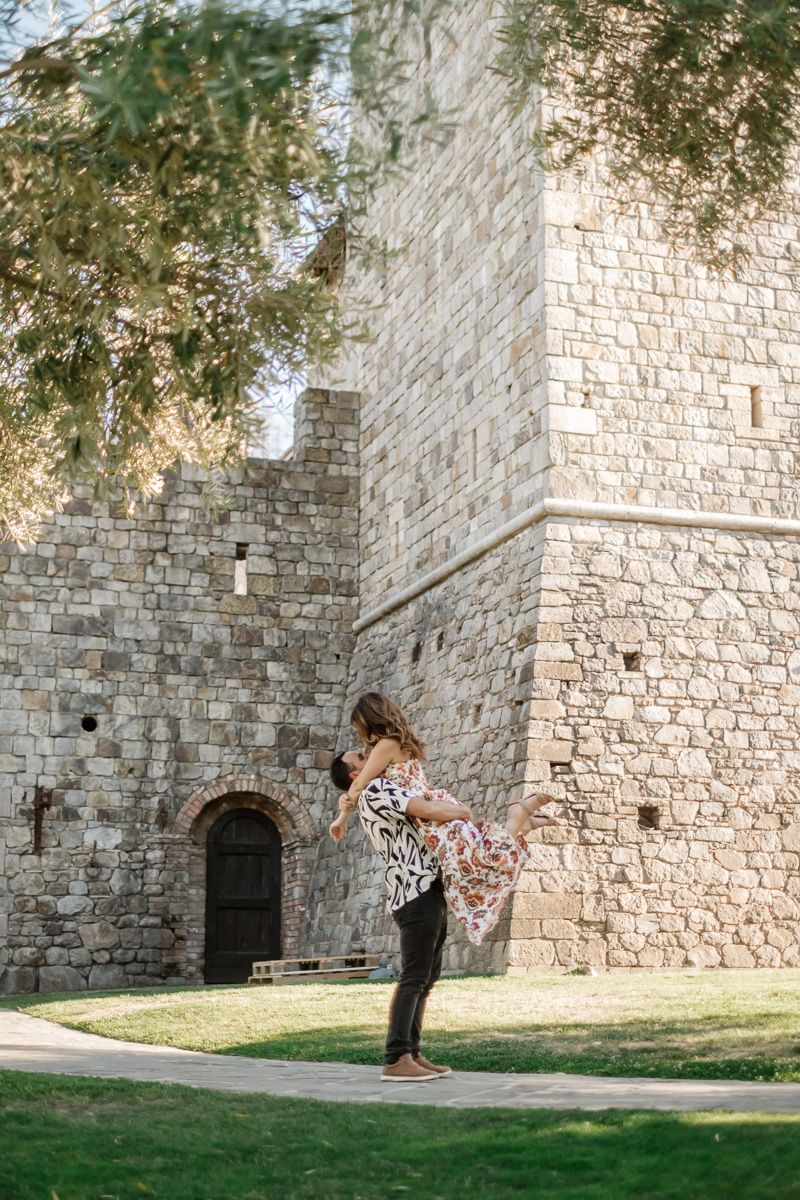 Couple embracing and dancing outdoors near a historic stone castle tower surrounded by lush green trees.