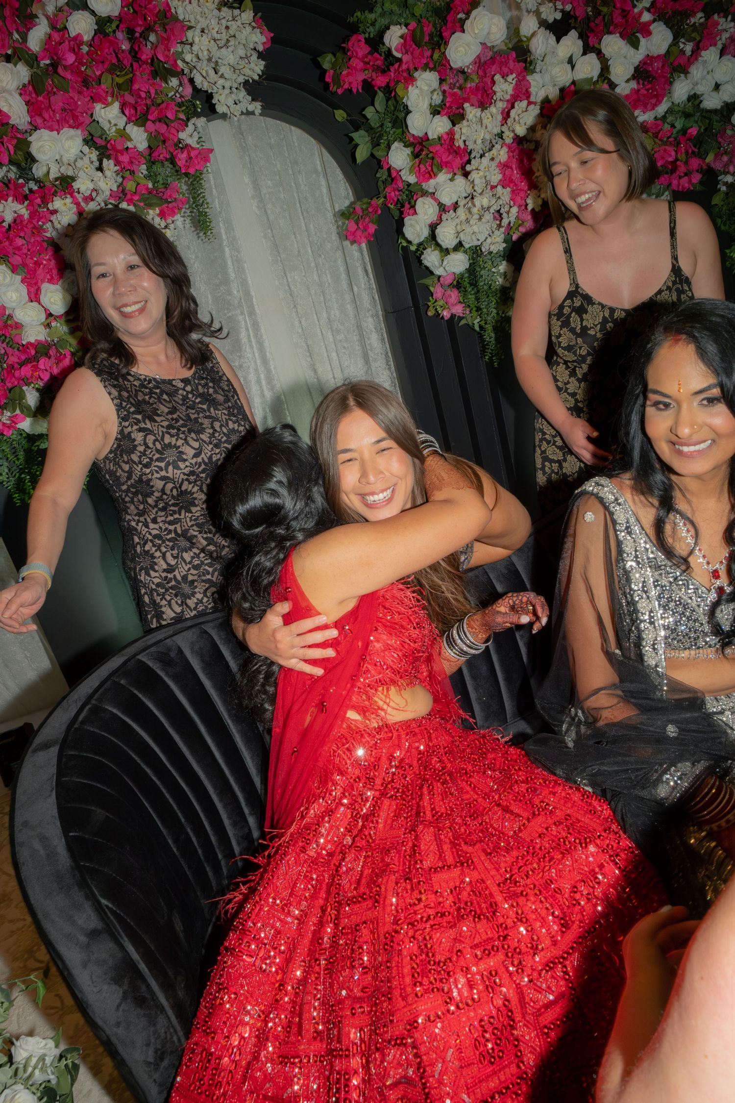 Joyful woman in red sequin saree hugs guest surrounded by friends near floral backdrop at reception.